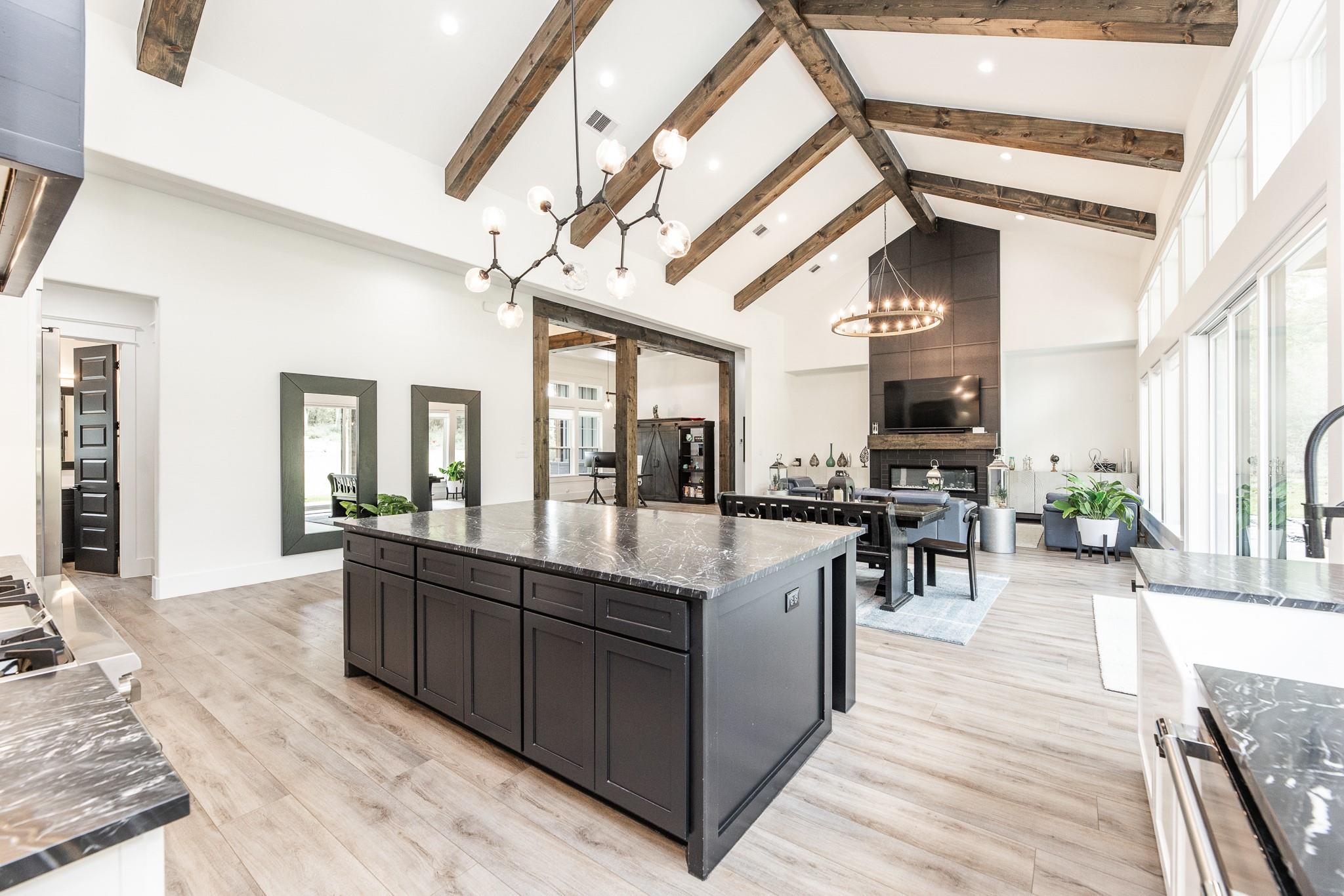 Open-plan kitchen with a large dark stone island, vaulted exposed-beam ceiling, and living area with fireplace.