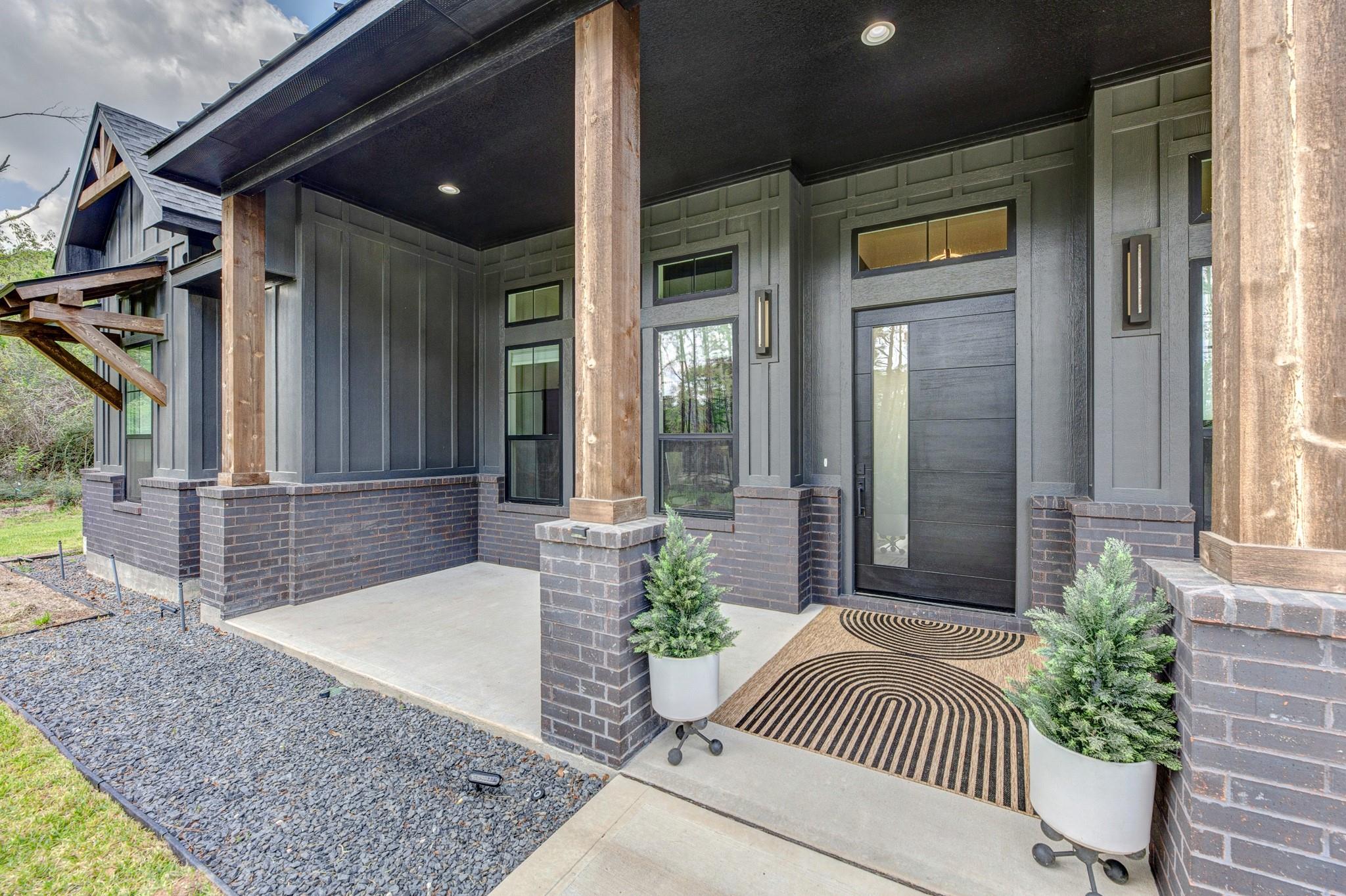 Modern dark-gray home’s covered front porch with a black door, timber posts, and potted evergreens.