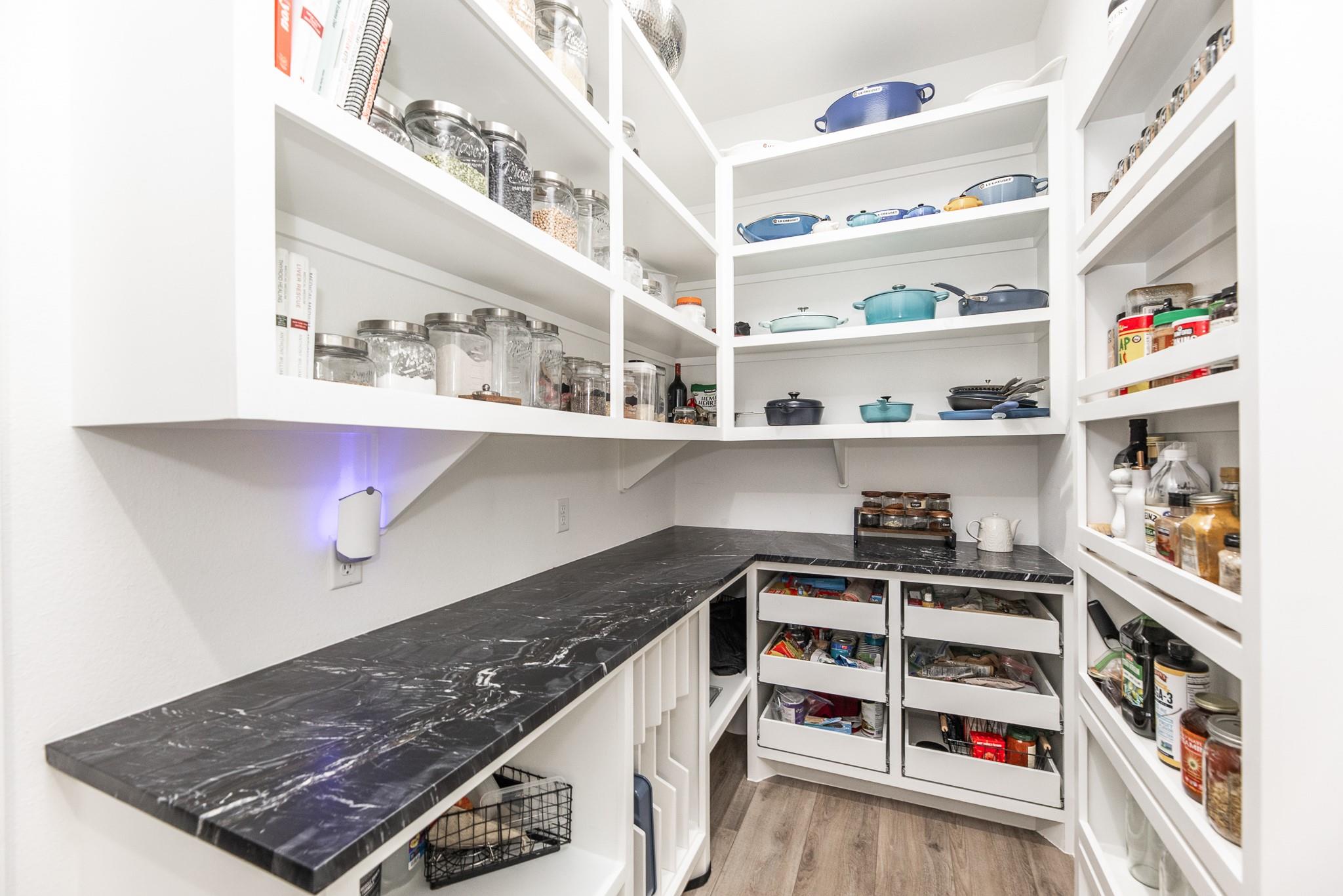 Walk-in pantry with open white shelving, black counters, and stocked pull-out drawers.