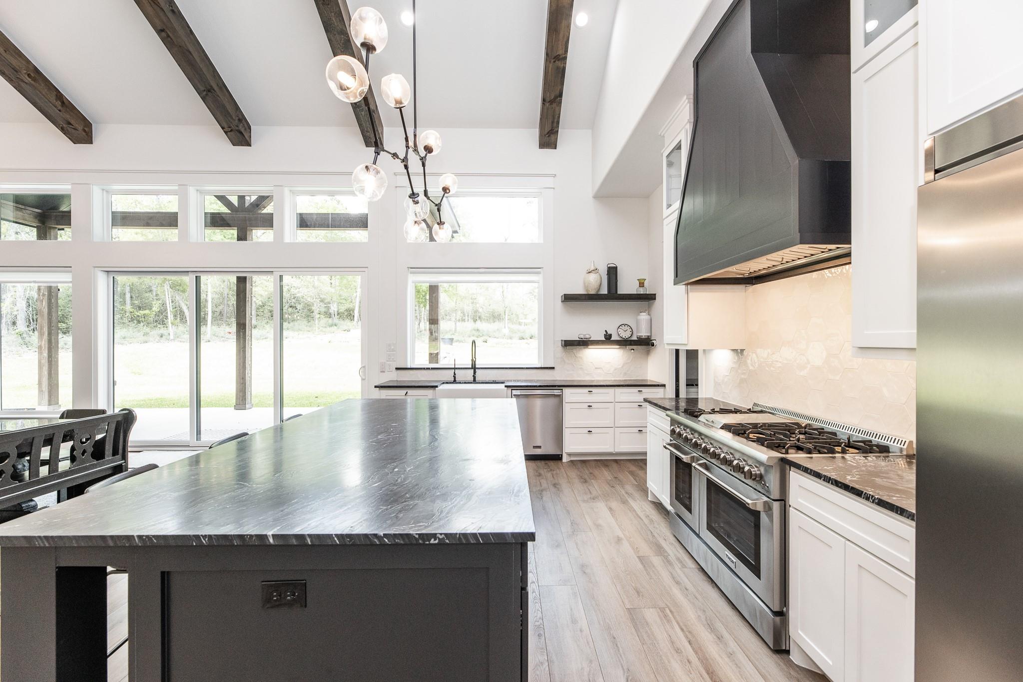 Bright kitchen with a large dark island, stainless steel range, exposed beams, and windows overlooking trees.