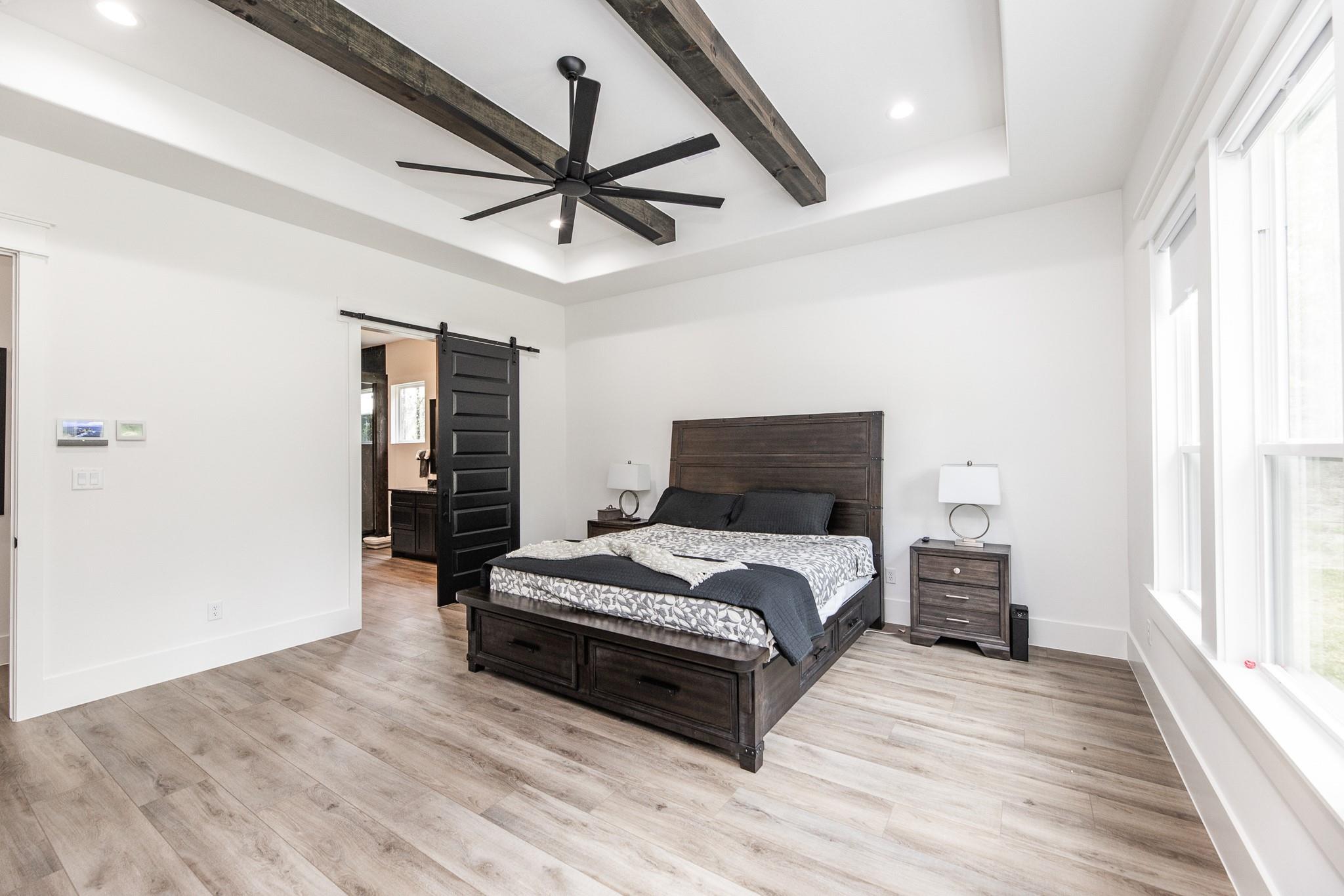 Furnished bedroom with dark wood bed, exposed ceiling beams, black fan, and tall windows.
