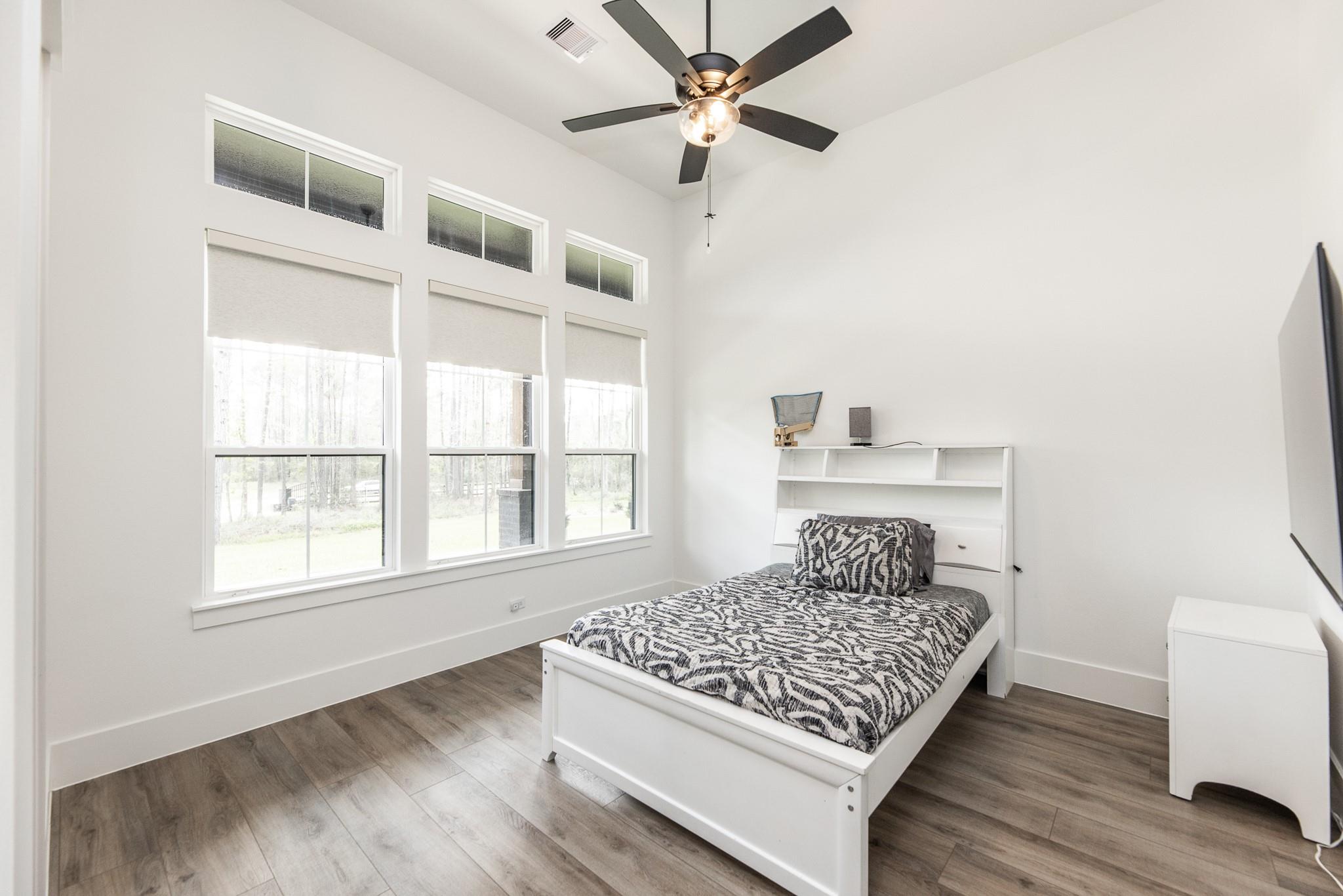 Bright bedroom with patterned bedding, three tall shaded windows, and a dark ceiling fan.