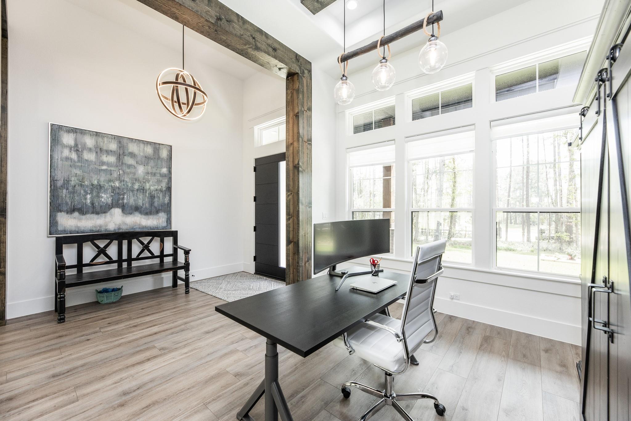 Bright home office with a black desk, white chair, large windows, and rustic wood beams.