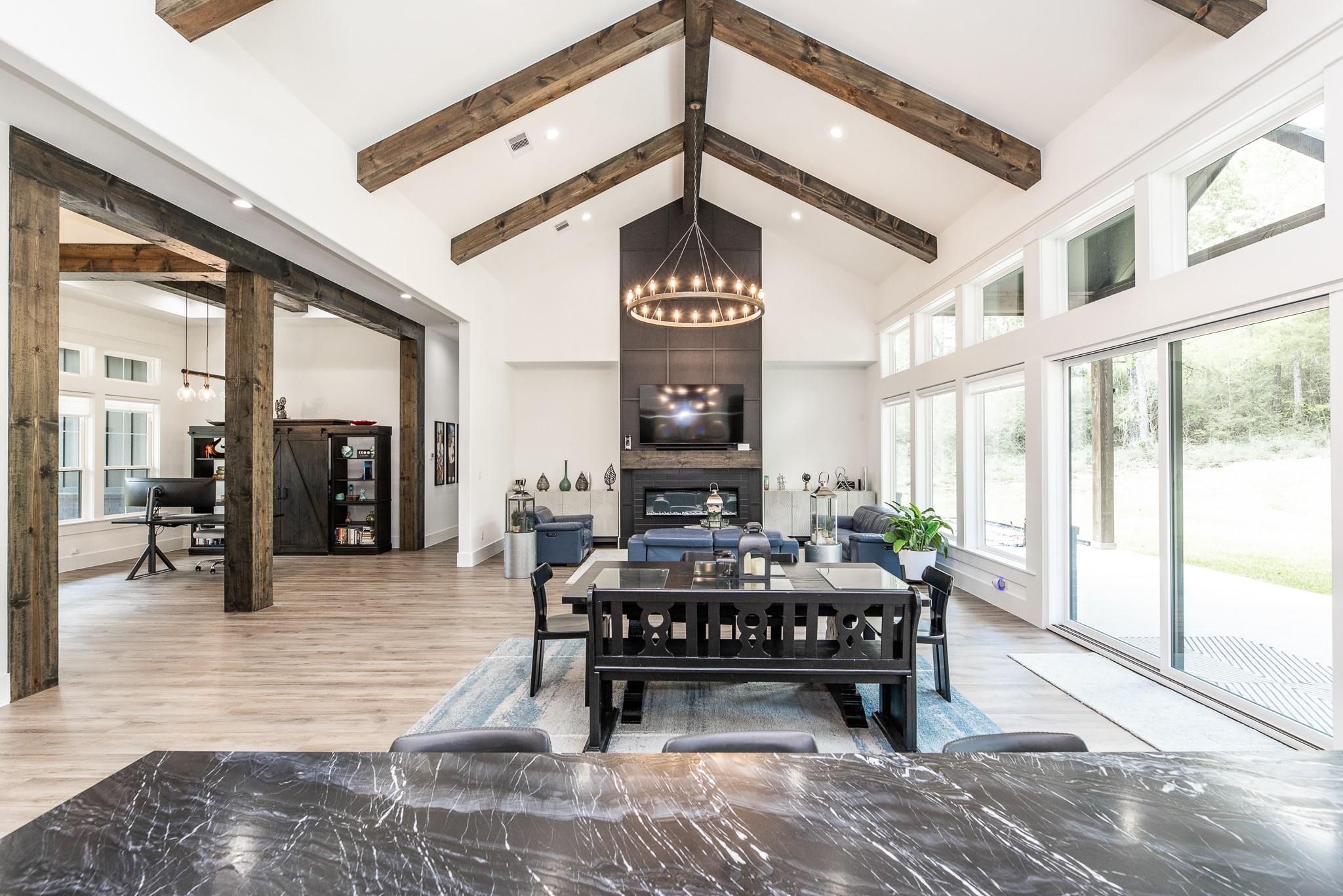 Open-plan living and dining room with vaulted wood-beam ceiling, fireplace, and tall windows overlooking a wooded yard.