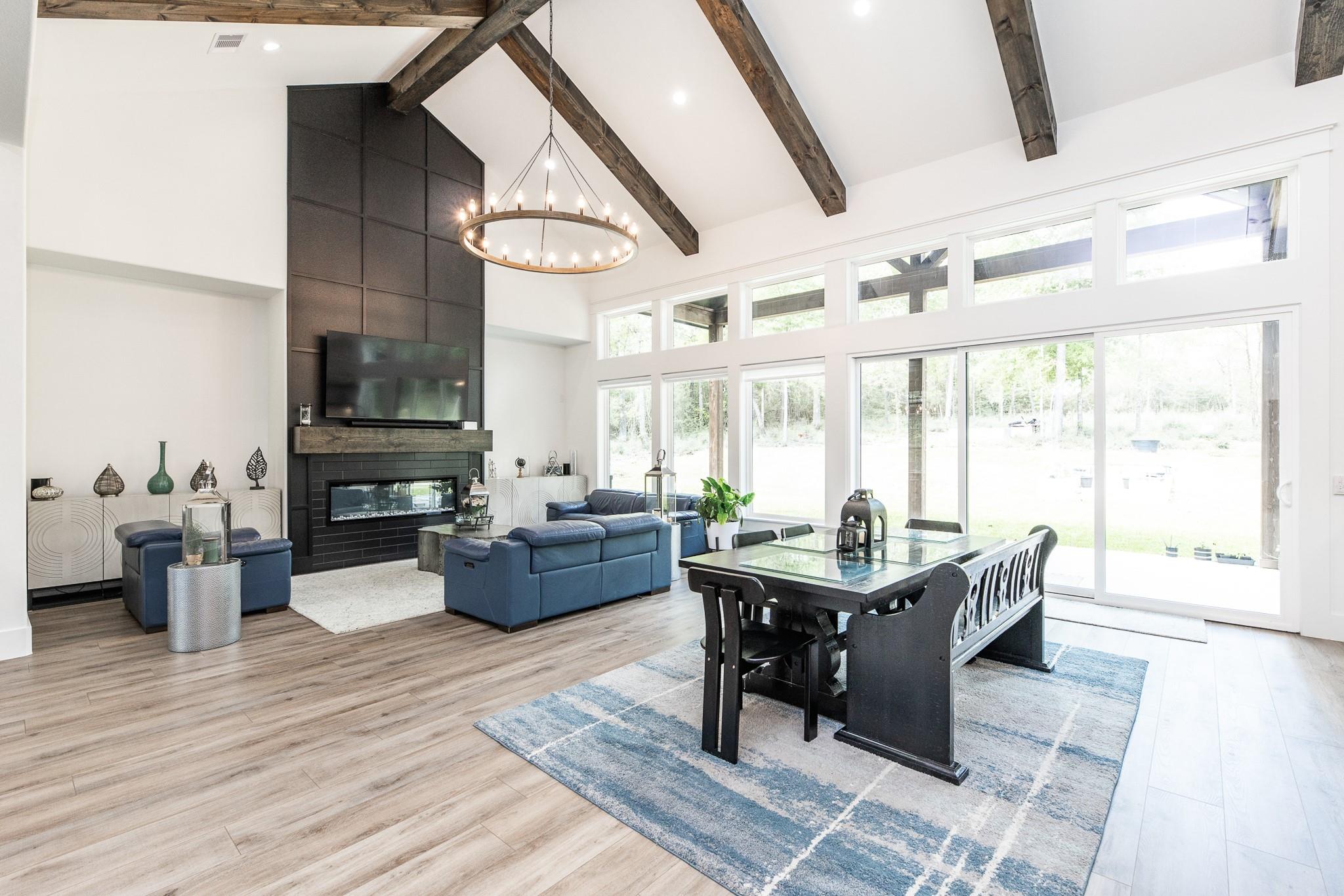 Open-plan living and dining room with exposed beams, a fireplace, blue seating, and large windows.