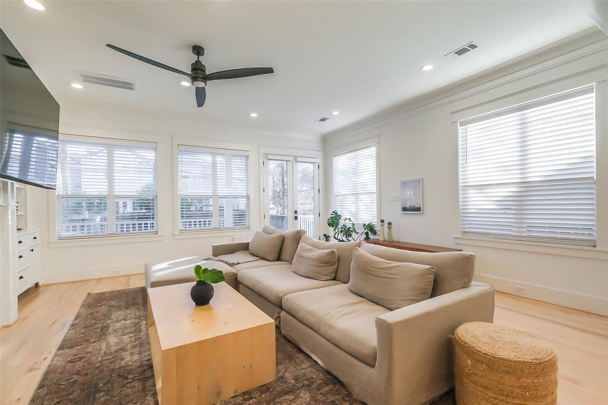 Bright living room with a beige sectional, light wood coffee table, and windows with white blinds.