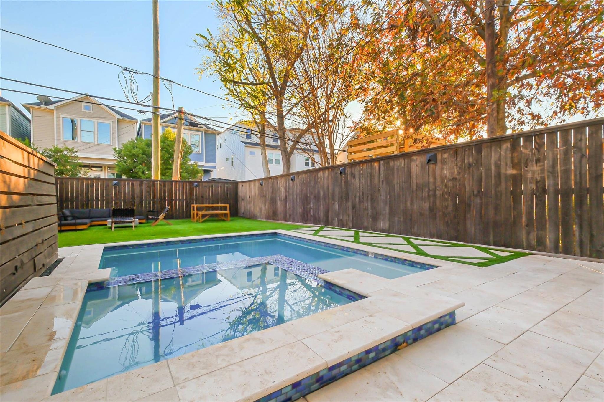 Backyard pool with pale stone decking, artificial turf, outdoor seating, and a tall wooden privacy fence.