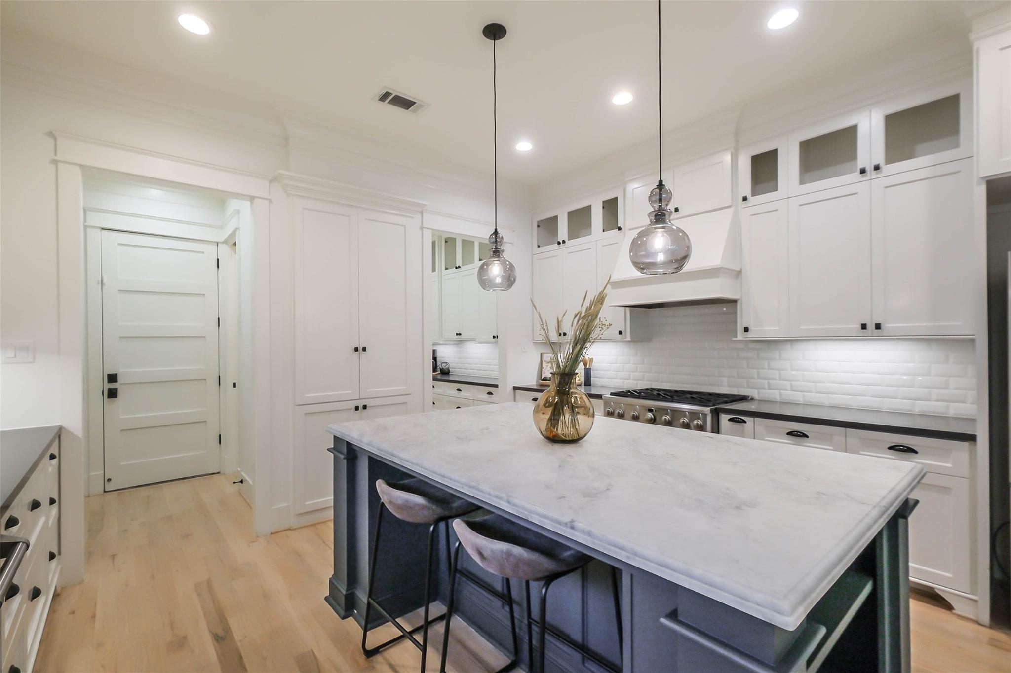 Bright kitchen with a large pale stone island, dark cabinetry, glass pendants, and white cabinets.