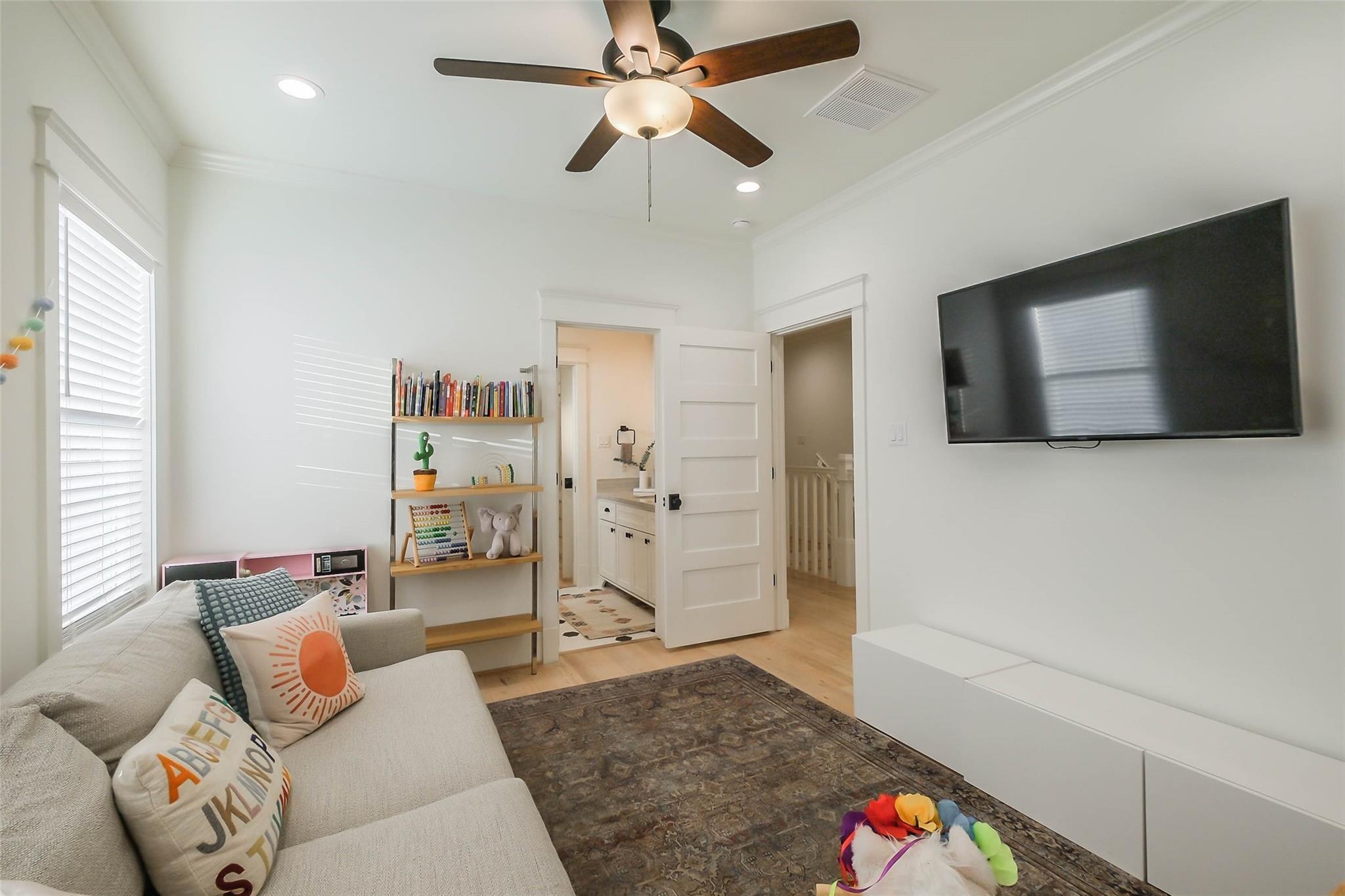 Living room with a sofa, wall-mounted TV, toy-filled bookshelf, and ceiling fan.