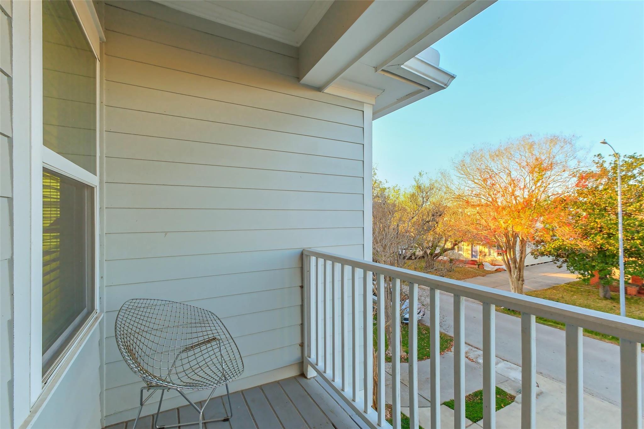 Covered balcony with a wire chair and white railing overlooking an autumn tree-lined street.