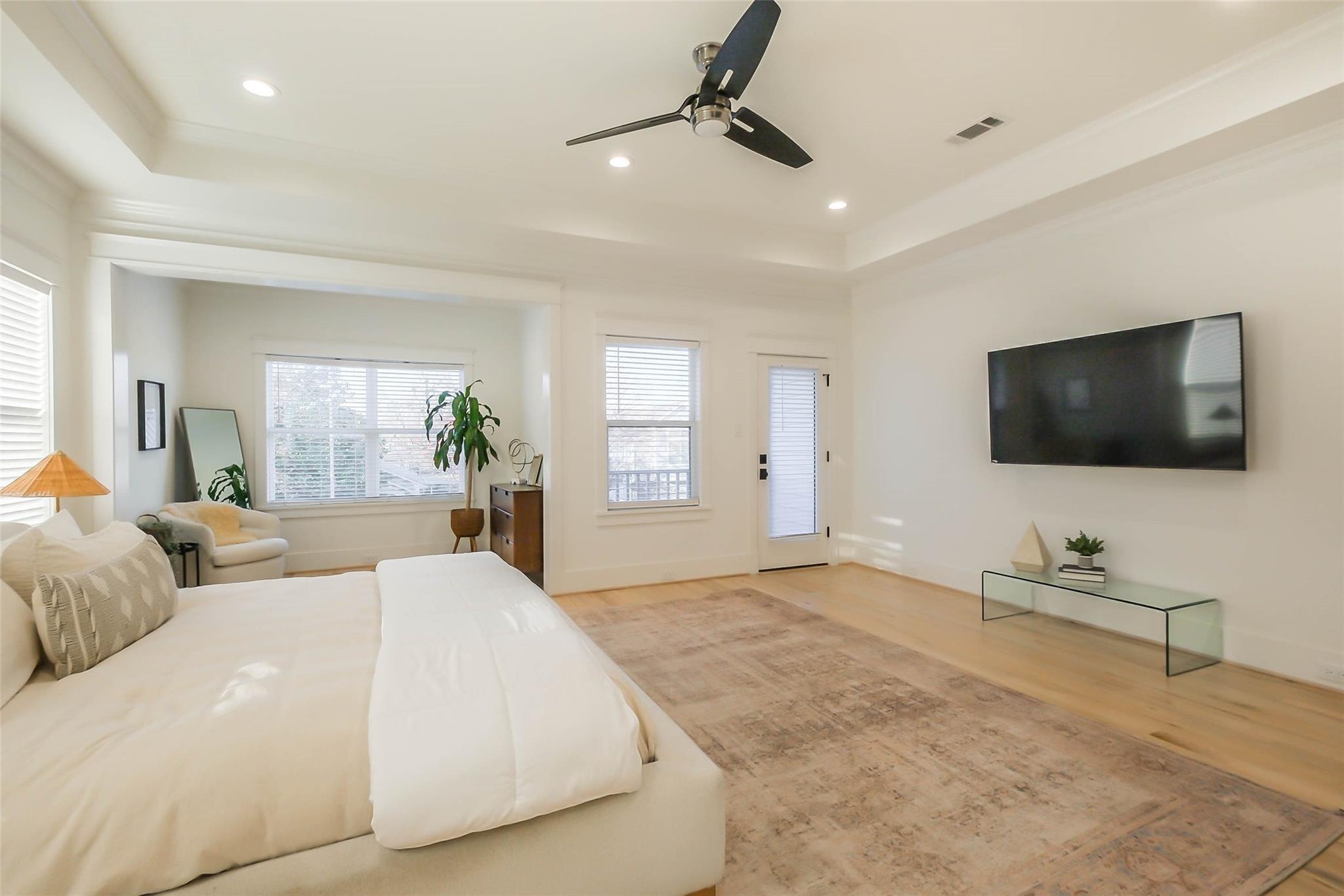 Bright bedroom with a large white bed, sitting area by shuttered windows, ceiling fan, and wall-mounted television.