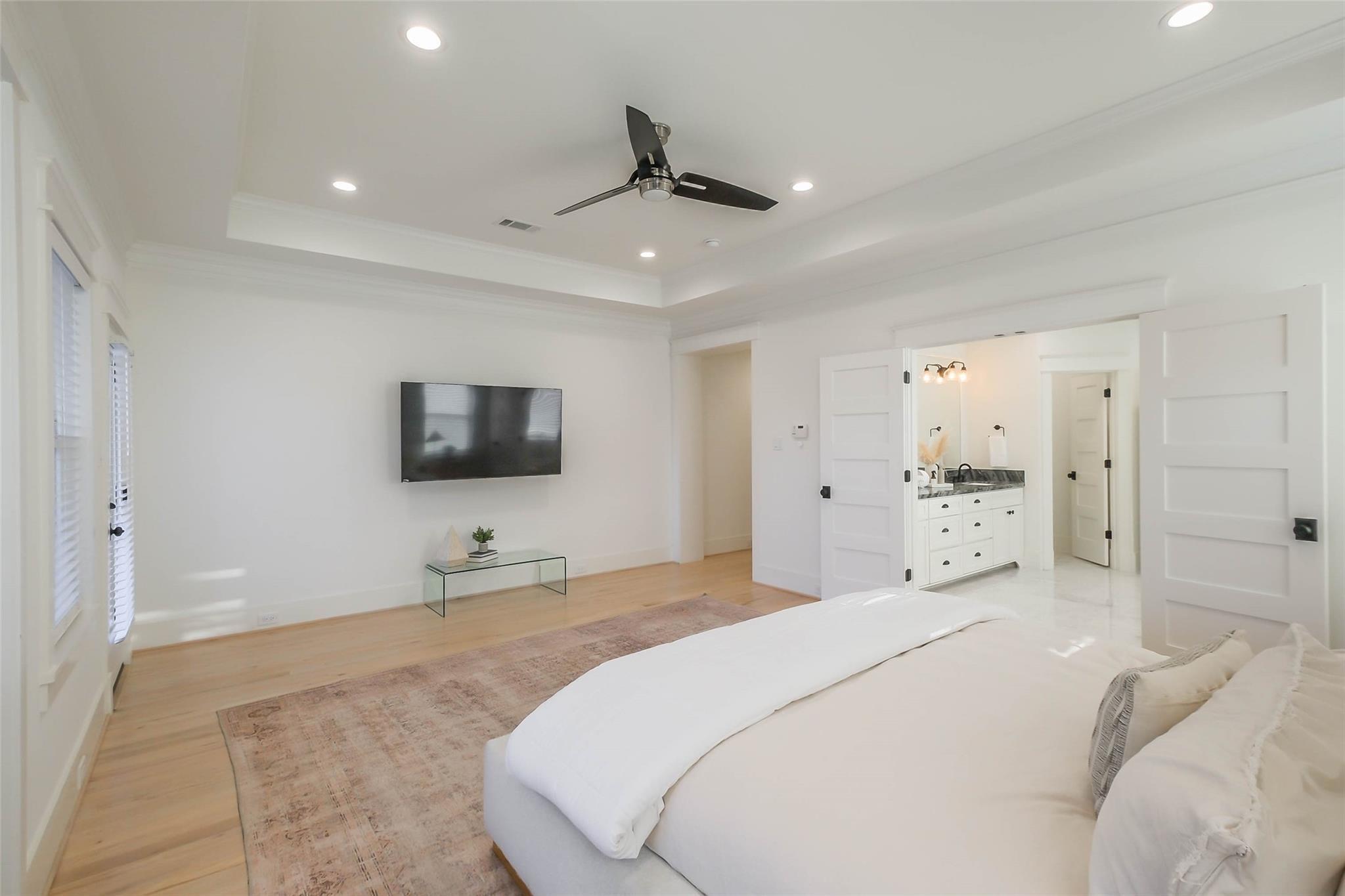 Bright staged bedroom with a large white bed, black ceiling fan, and open doors to an adjoining bathroom.