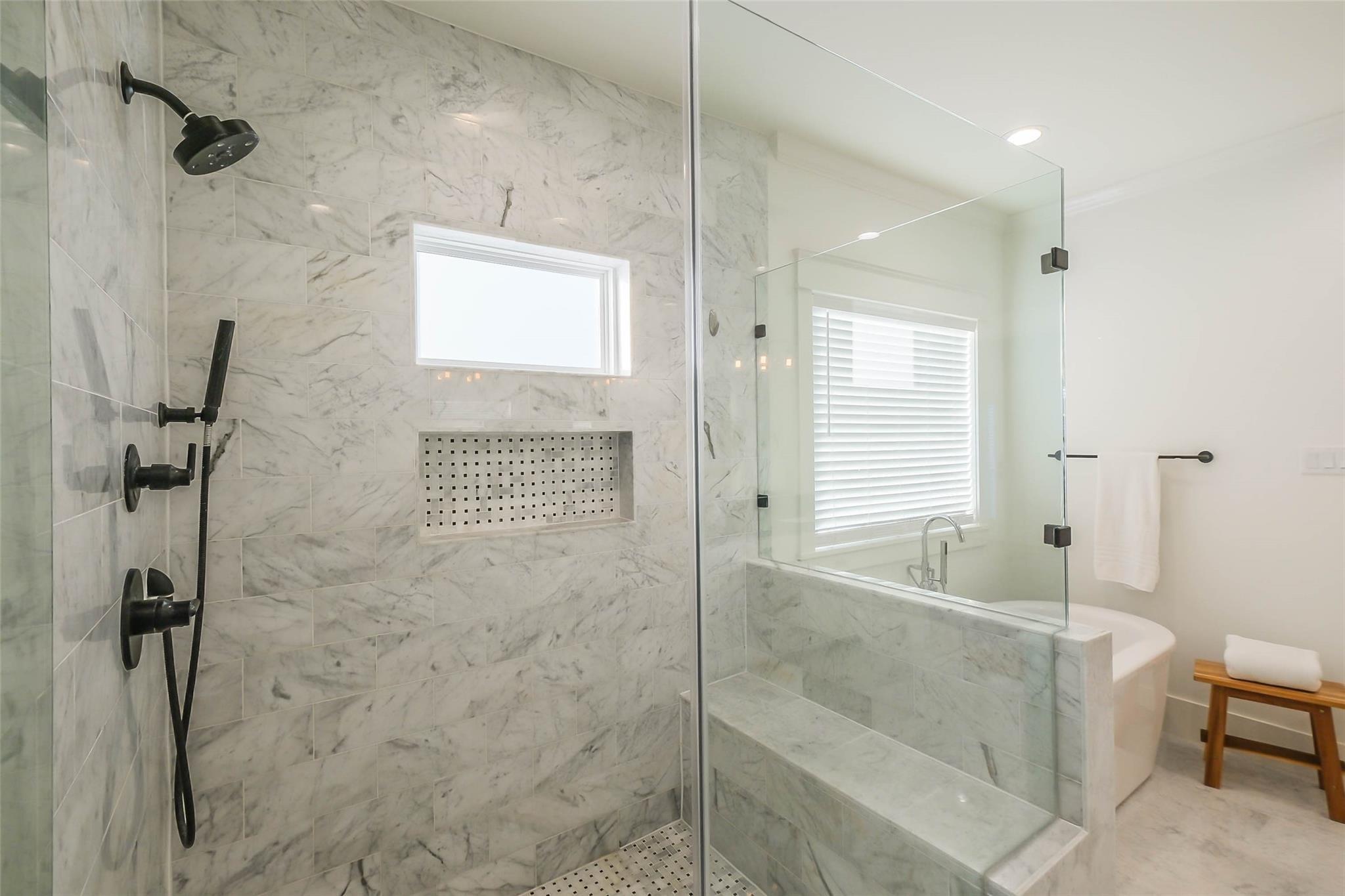 Marble-tiled walk-in shower with black fixtures beside a bathtub and window.