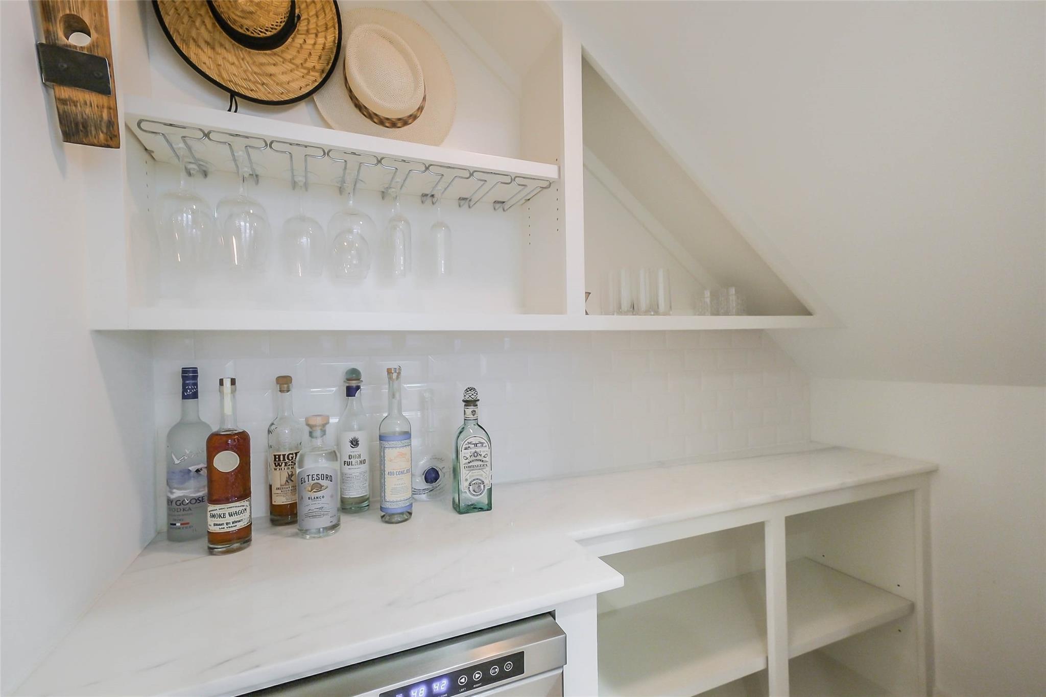 Built-in white bar with liquor bottles, hanging glasses, and an under-counter beverage refrigerator.