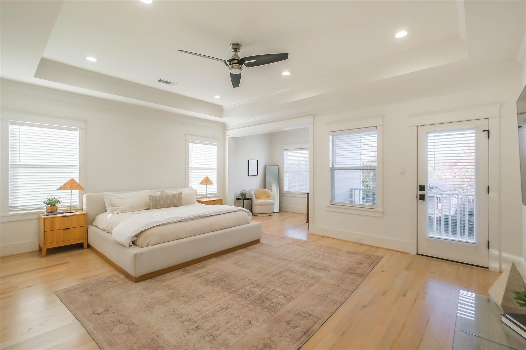 Bright staged bedroom with a large upholstered bed, pale wood floors, and a ceiling fan.