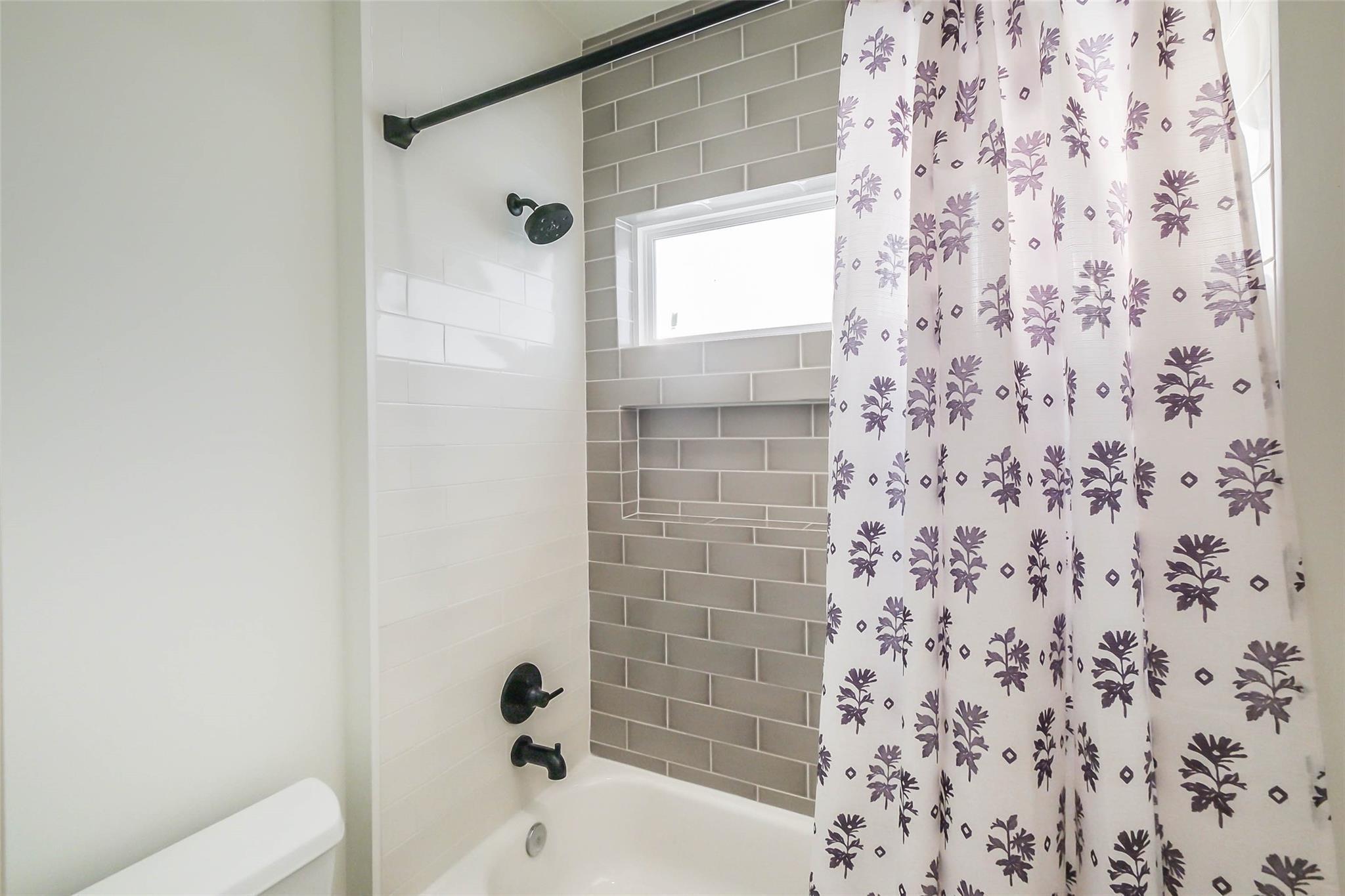 Bathroom tub and shower with gray subway tile, a recessed niche, black fixtures, and a purple floral curtain.