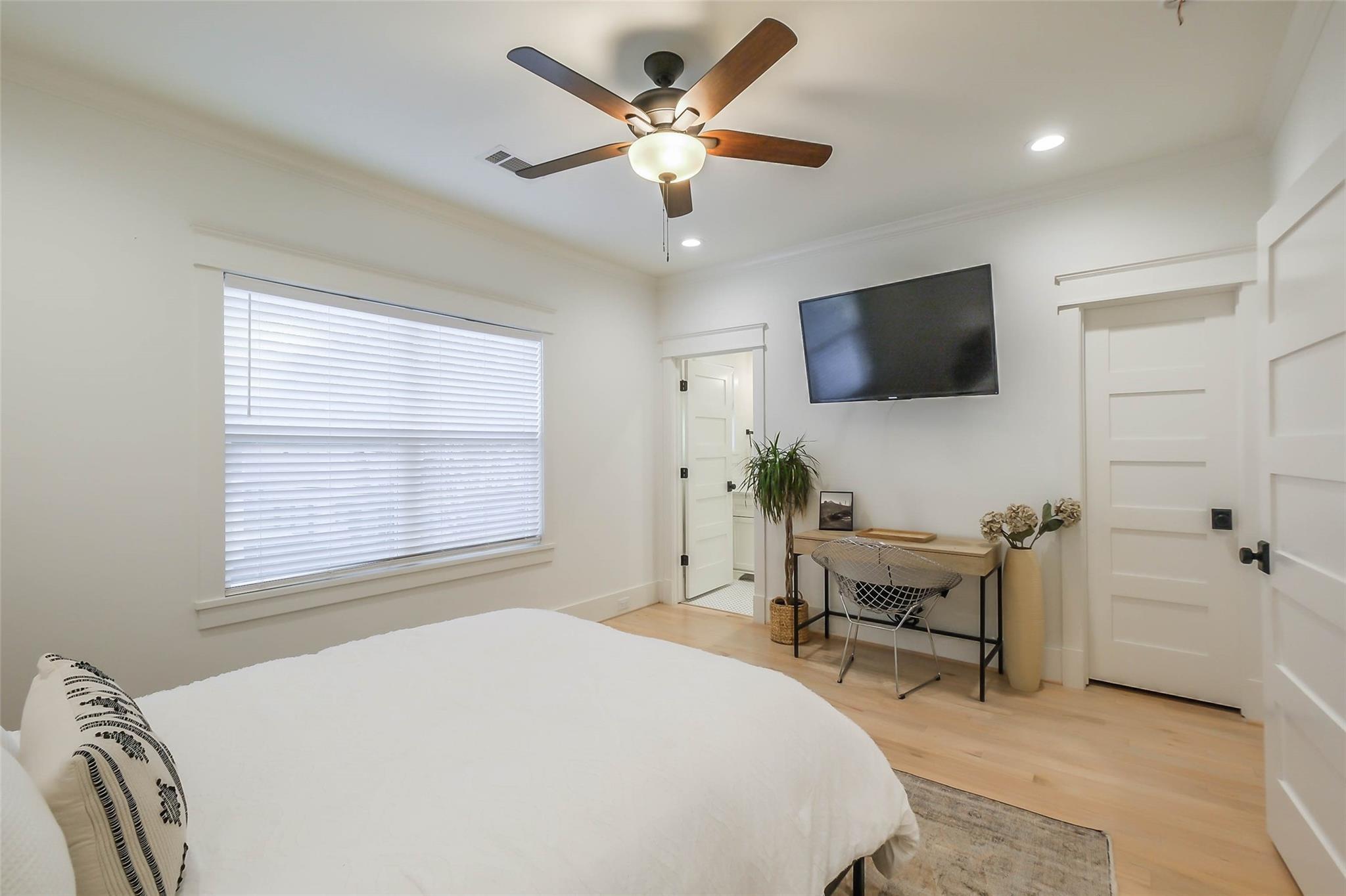 Bright bedroom with a white bed, ceiling fan, wall-mounted television, and window with blinds.