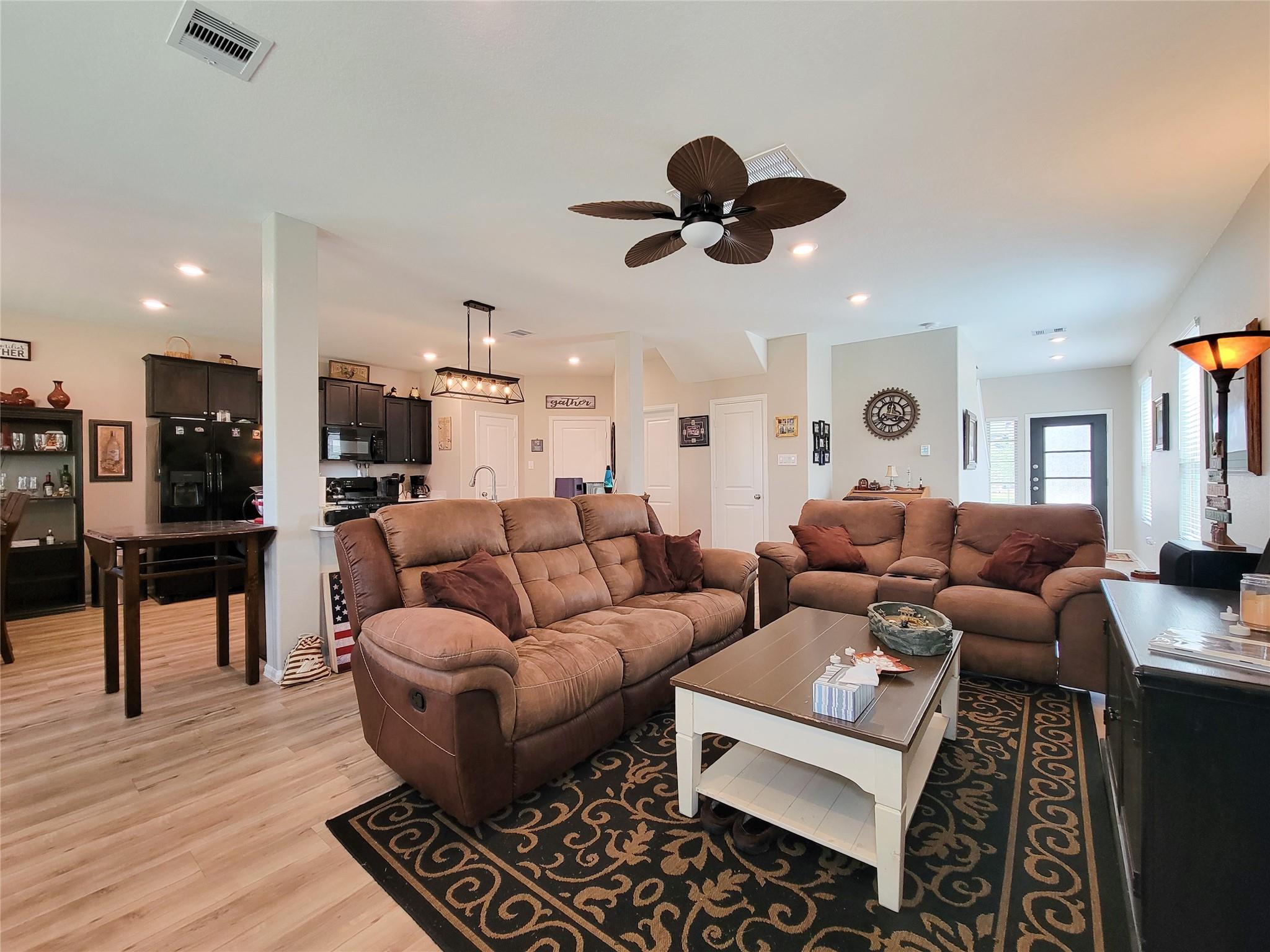 Open-plan living room with brown reclining sofas, patterned rug, ceiling fan, and dark-cabinet kitchen.
