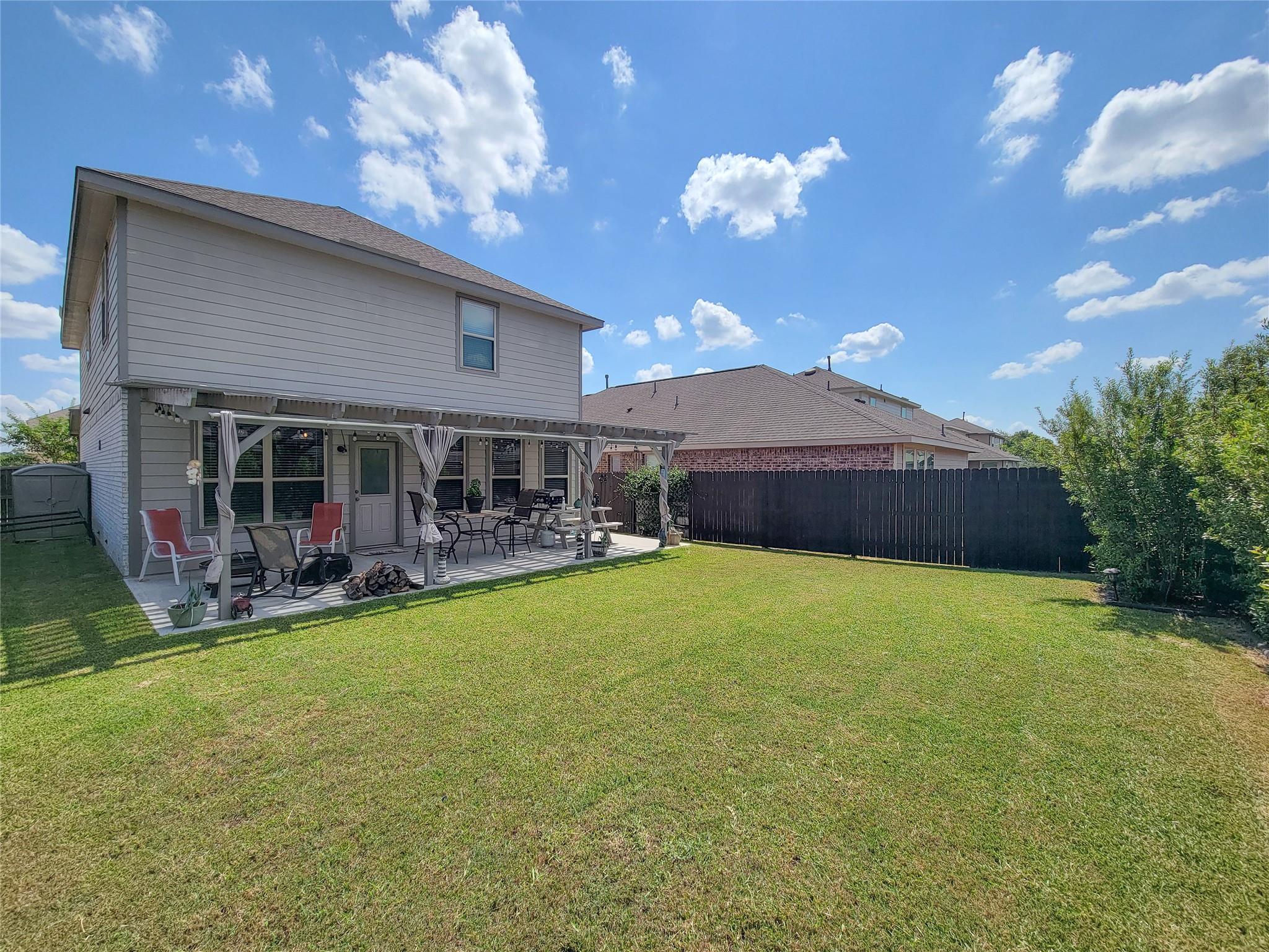 Two-story home with a covered, furnished patio overlooking a broad fenced backyard lawn.