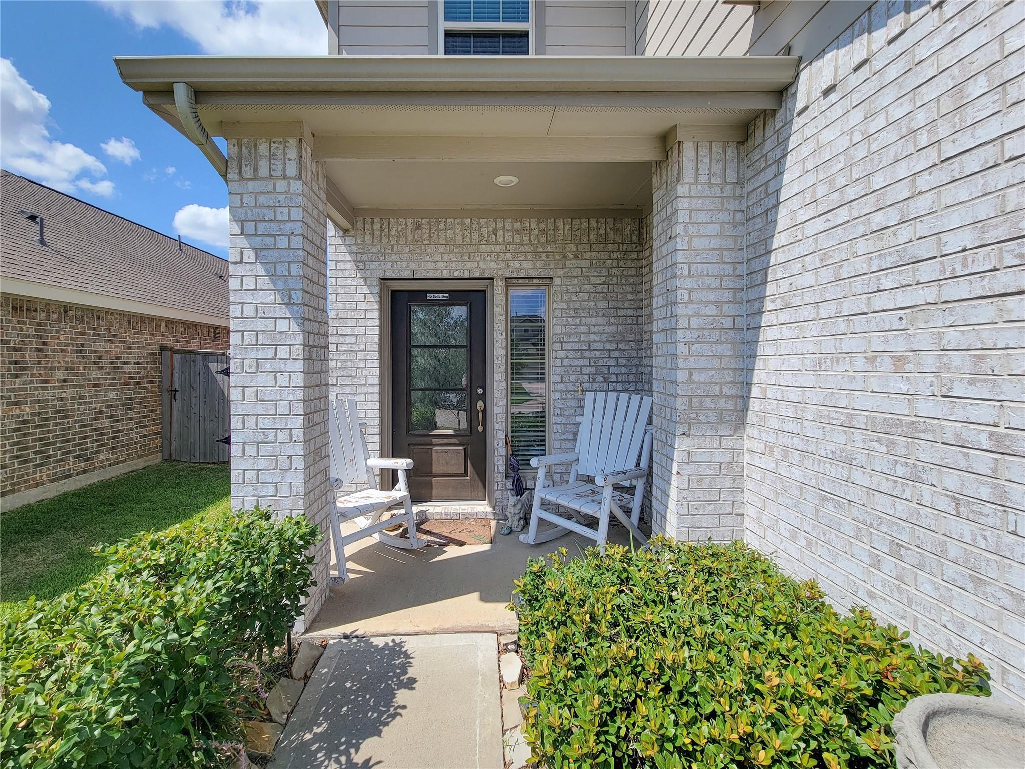 Covered brick front porch with a dark entry door, white rocking chairs, and trimmed hedges.