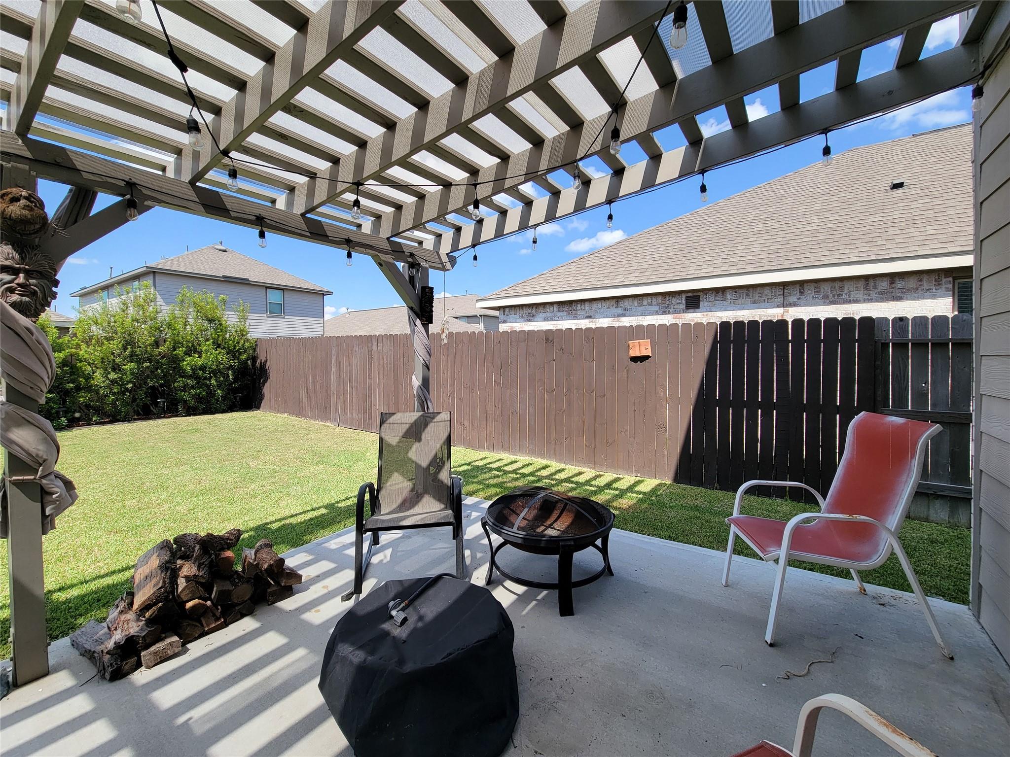 Backyard patio beneath a string-lit pergola, with a fire pit, chairs, and fenced lawn.