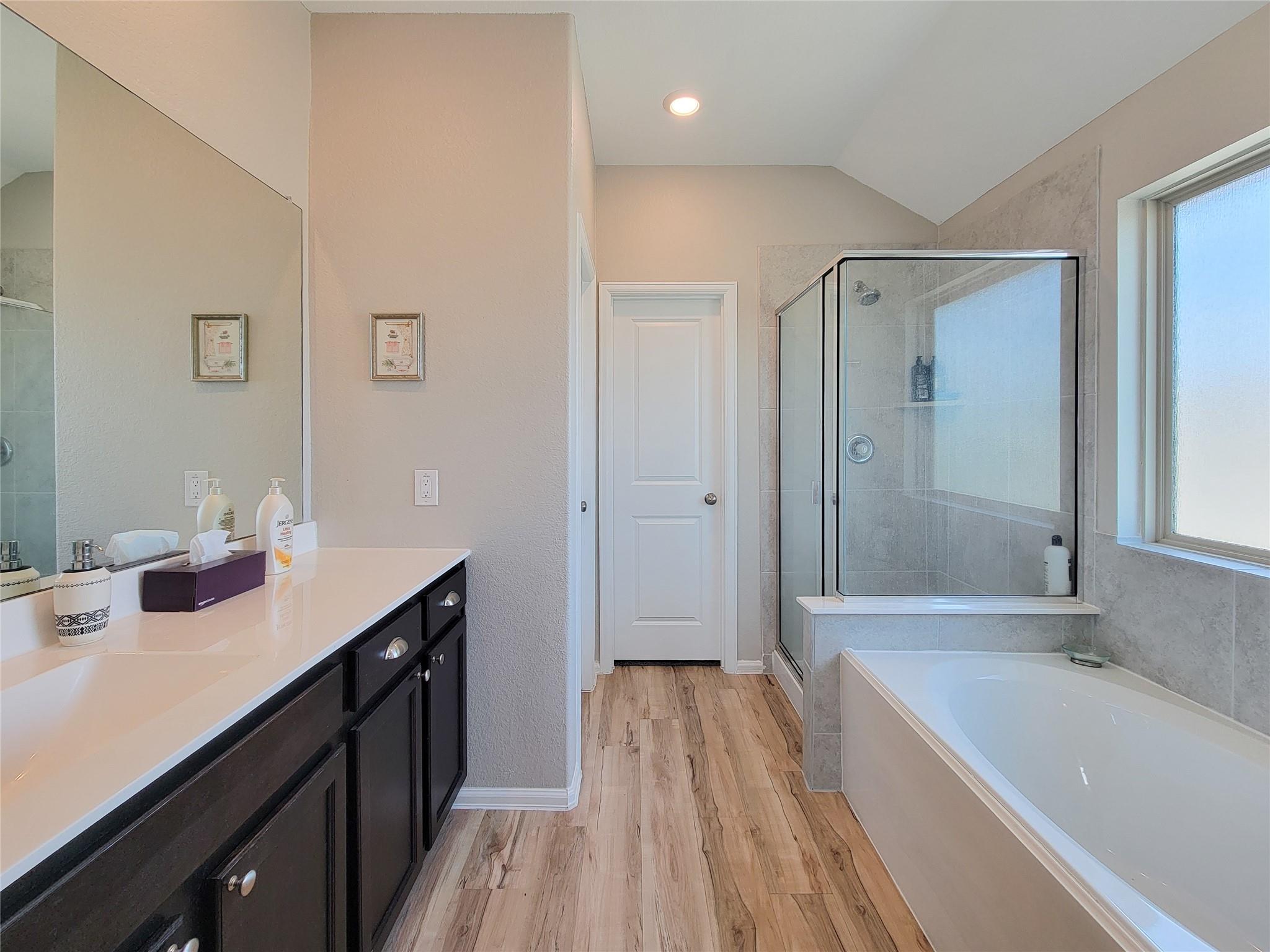 Bathroom with a glass-enclosed shower, built-in tub, dark vanity, and wood-look flooring.