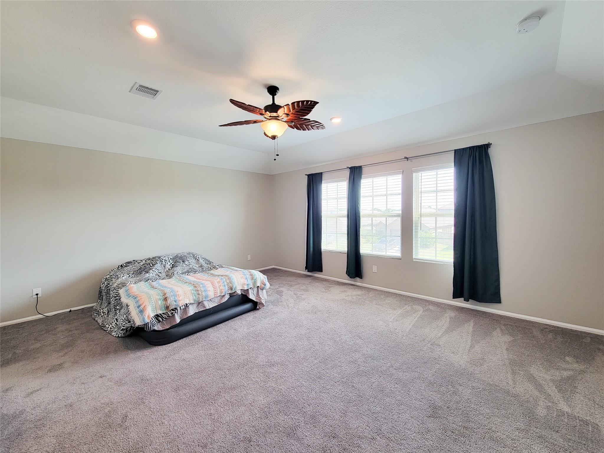 Spacious carpeted bedroom with a ceiling fan, three curtained windows, and a bed.