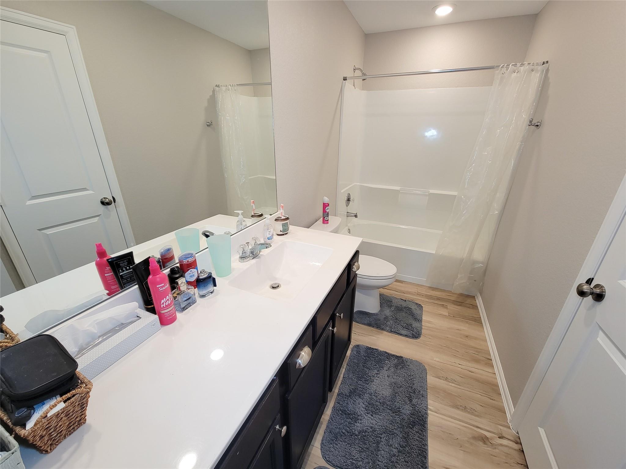 Bathroom with a dark vanity, large mirror, tub-shower, toilet, and wood-look flooring.