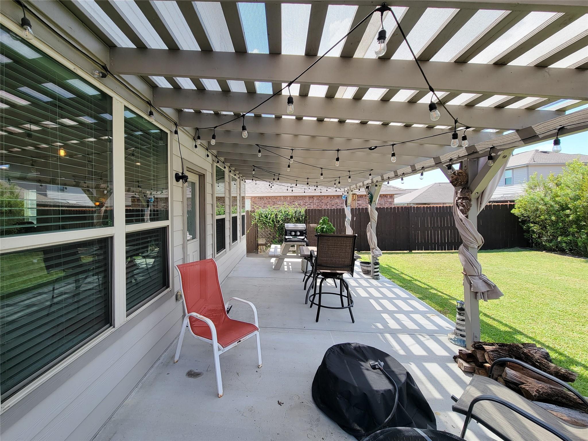 Covered backyard patio with pergola, string lights, outdoor seating, and grill beside a fenced lawn.