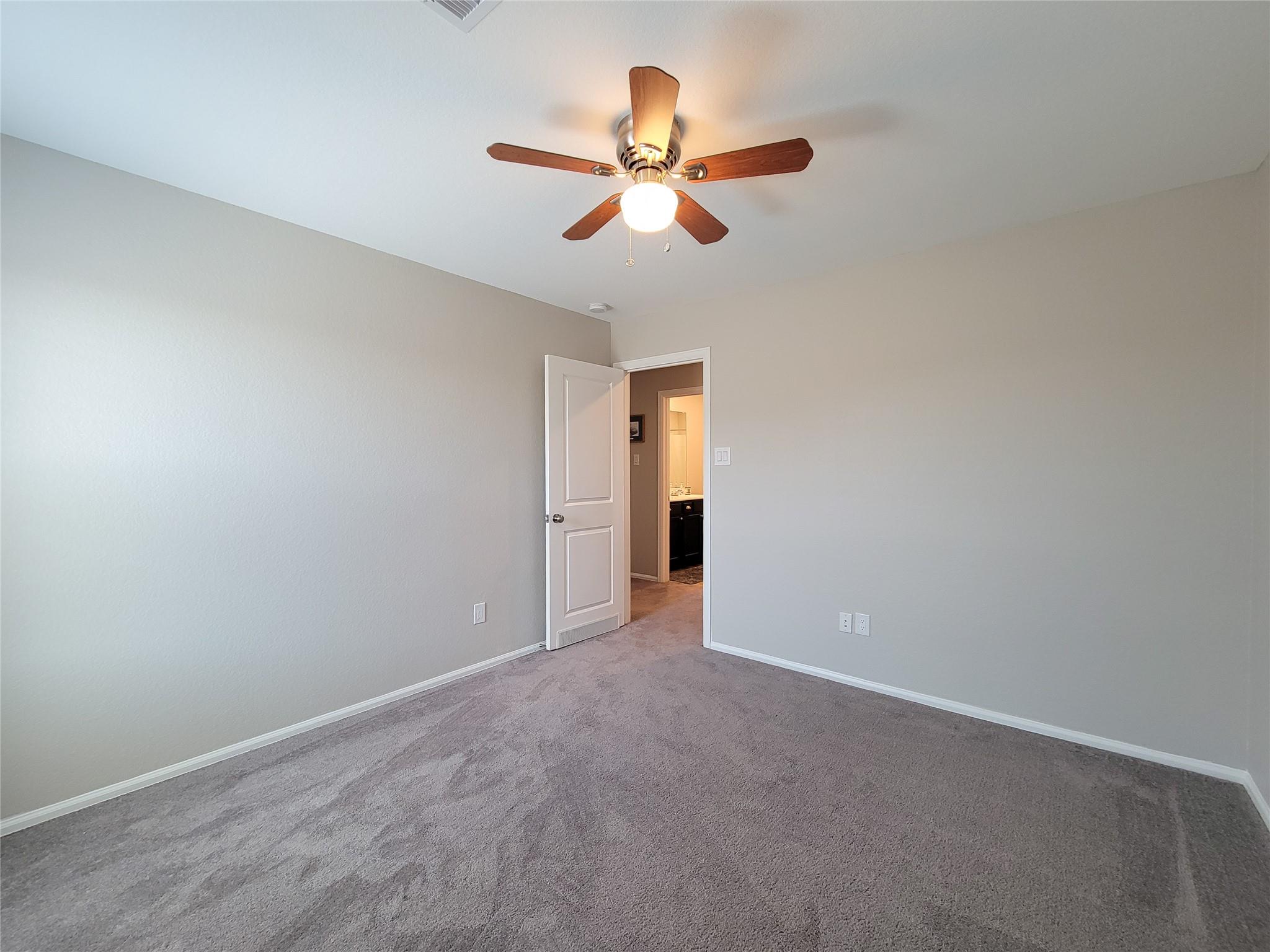 Empty bedroom with gray carpet and a ceiling fan, viewed toward an open bathroom door.