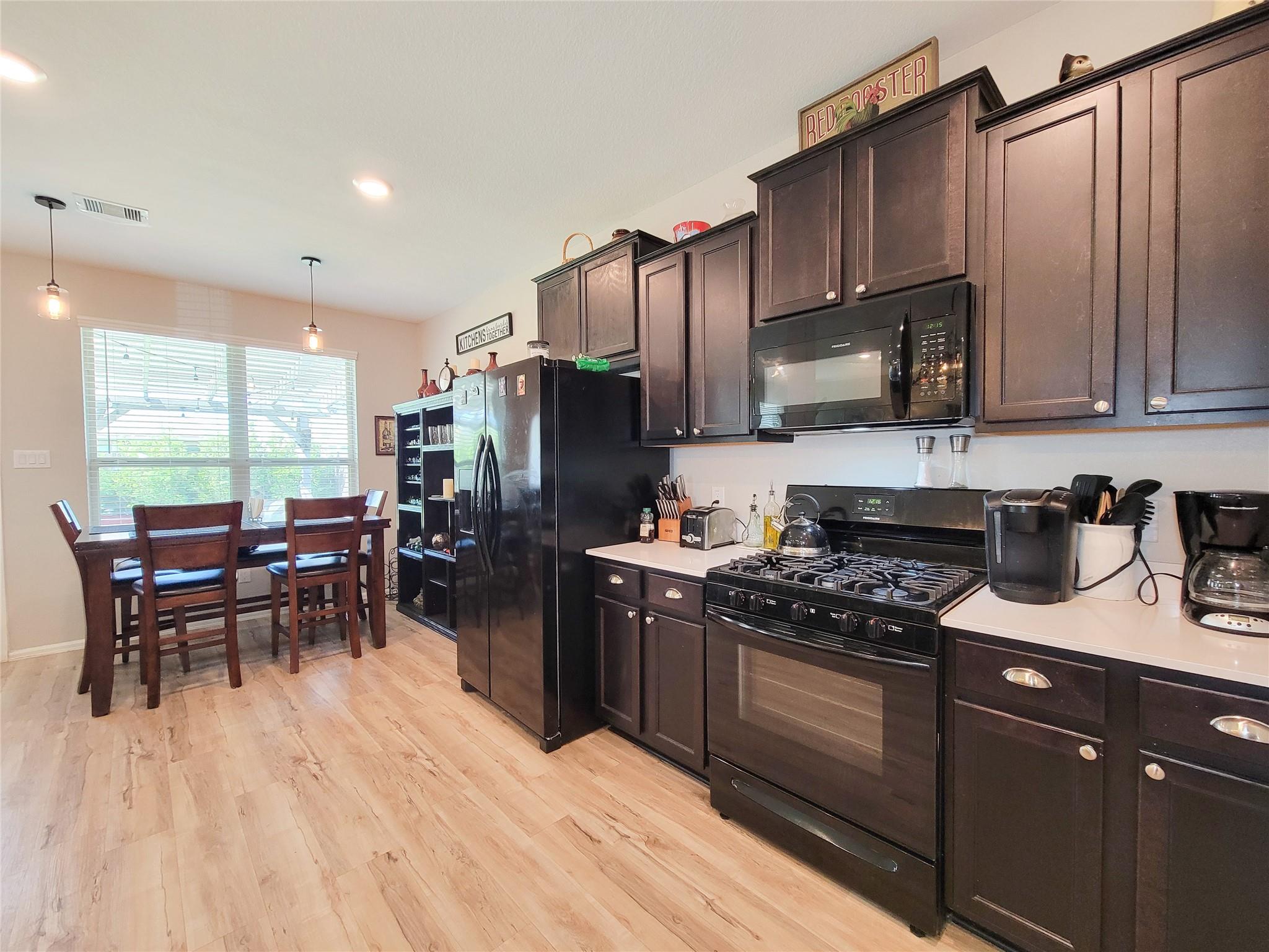Kitchen with dark cabinets and black appliances, opening to a dining area by a large window.