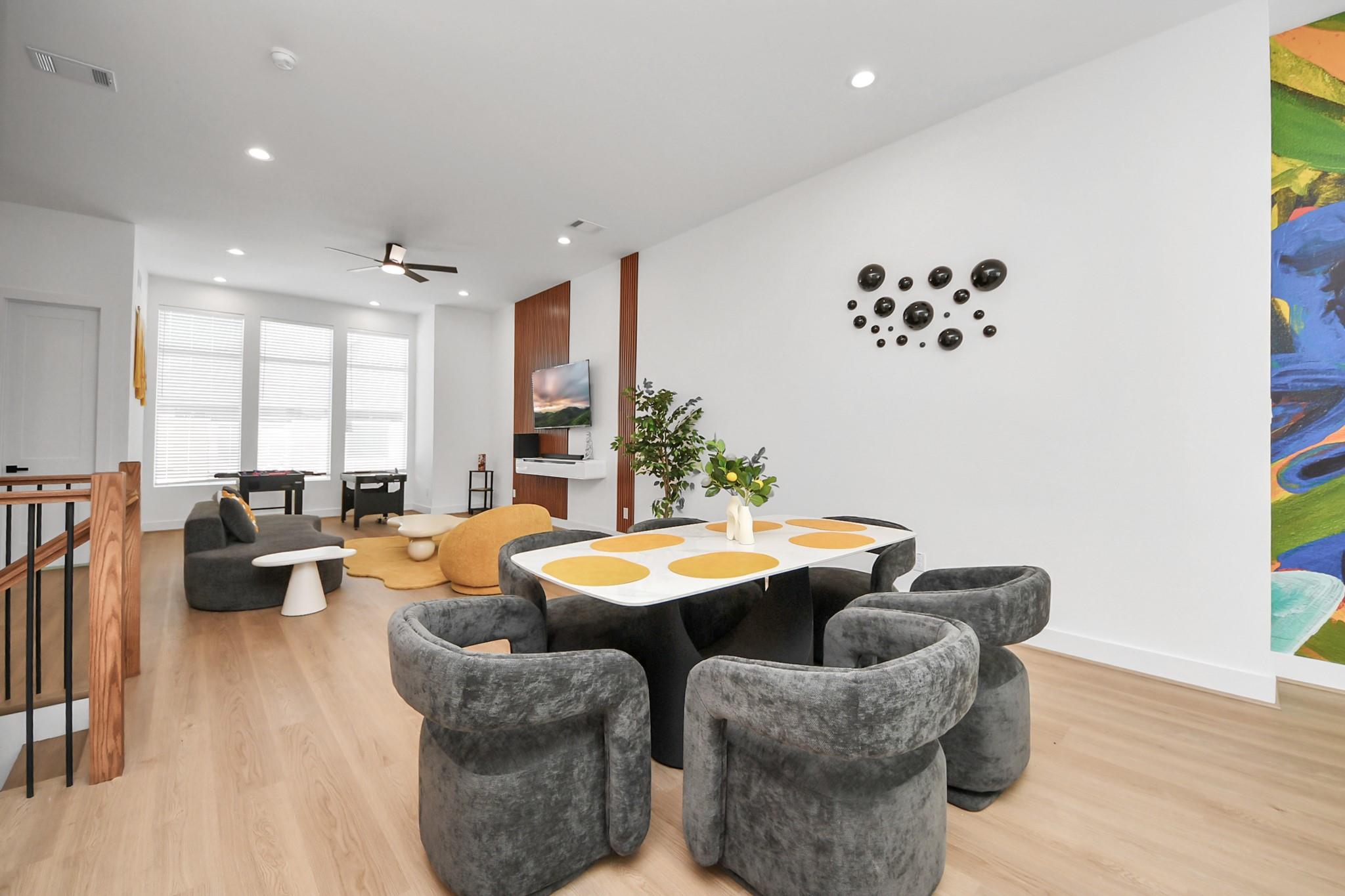 Open-plan dining and living area with rounded gray chairs, light wood floors, and a lounge with game tables.