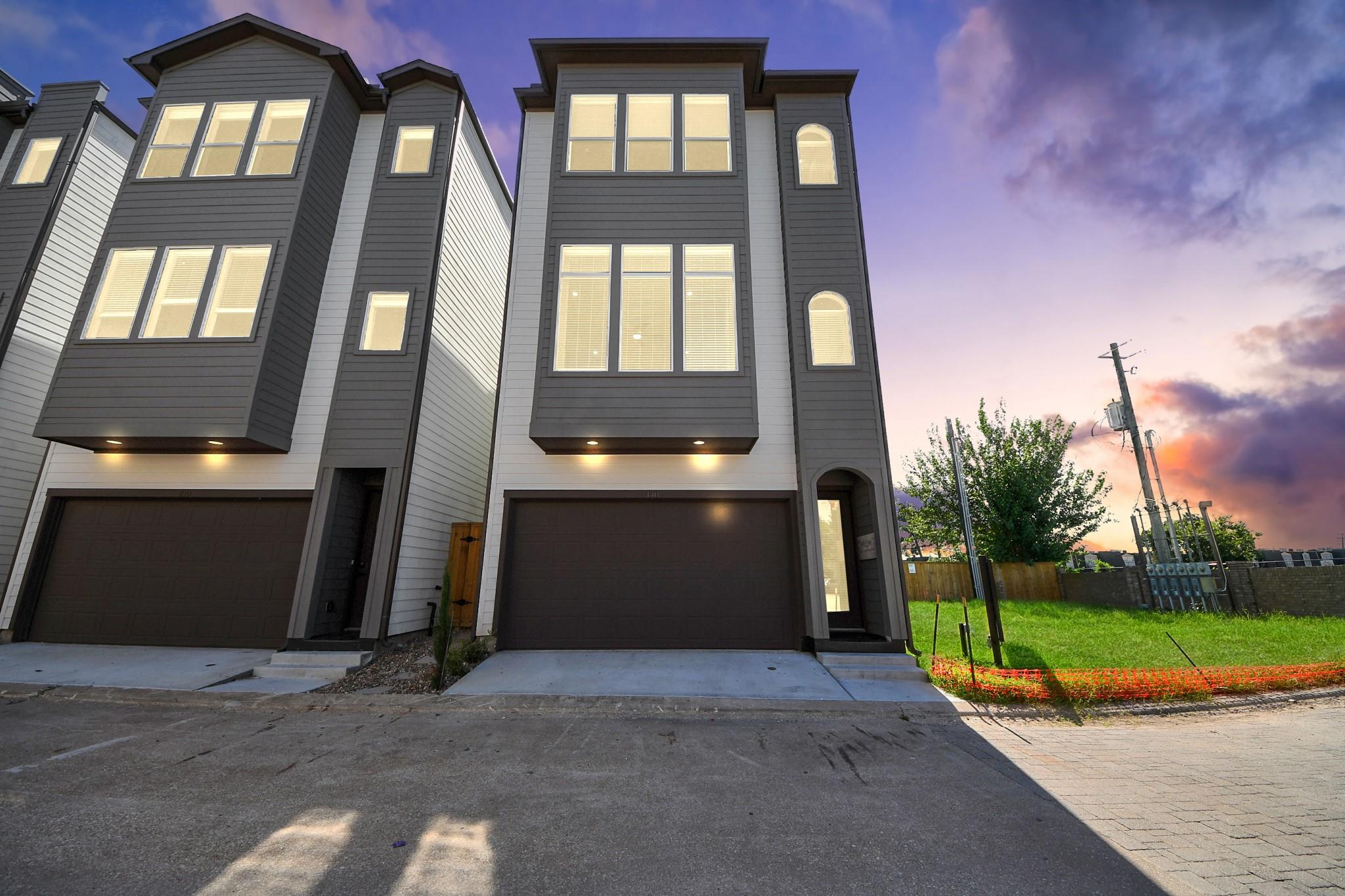 Modern gray townhome with illuminated windows and a dark garage door at dusk.