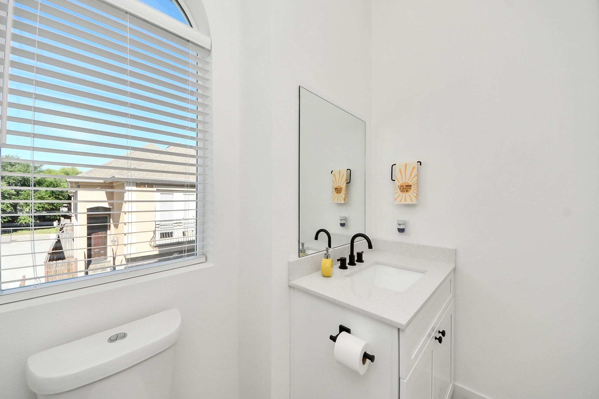 Bright white bathroom with a black faucet, mirrored wall, and window with horizontal blinds.