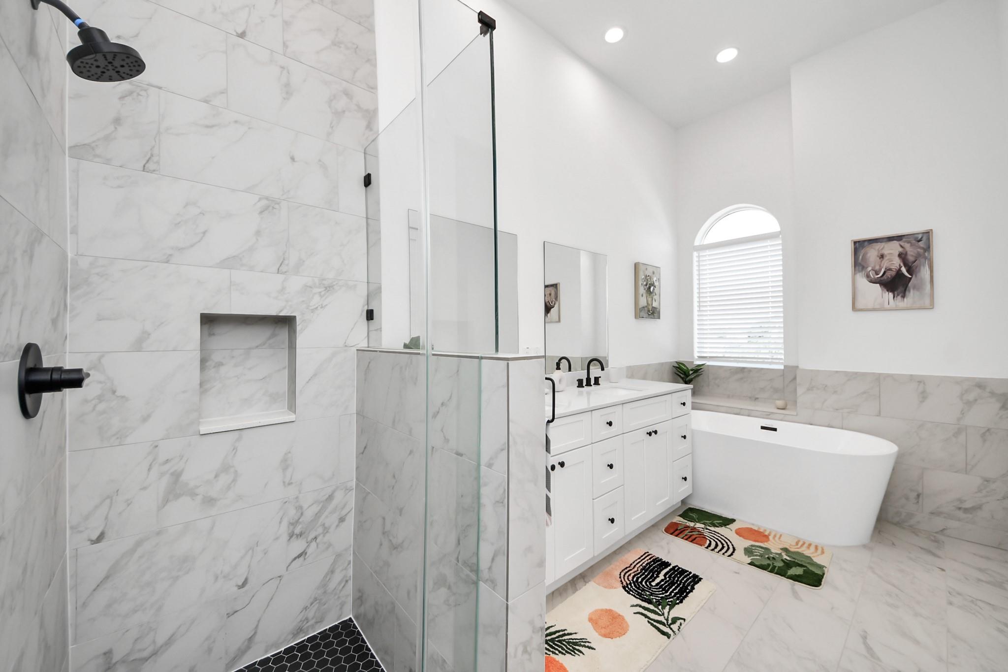 Bright marble-tiled bathroom with glass shower, freestanding tub, and white vanity with black fixtures.