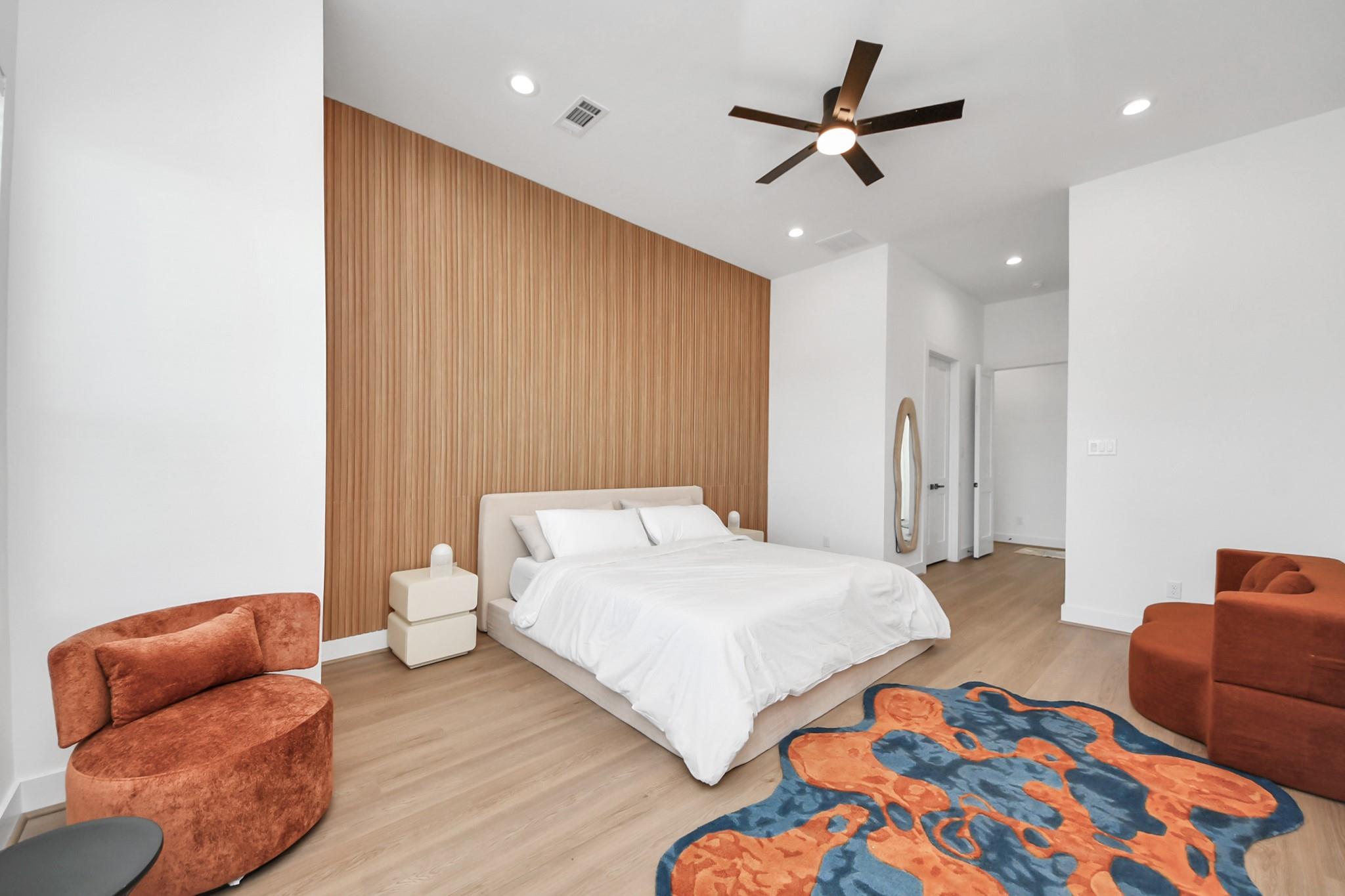 Staged bedroom with a white bed, wood-slat accent wall, rust-colored chairs, and a blue-and-orange rug.