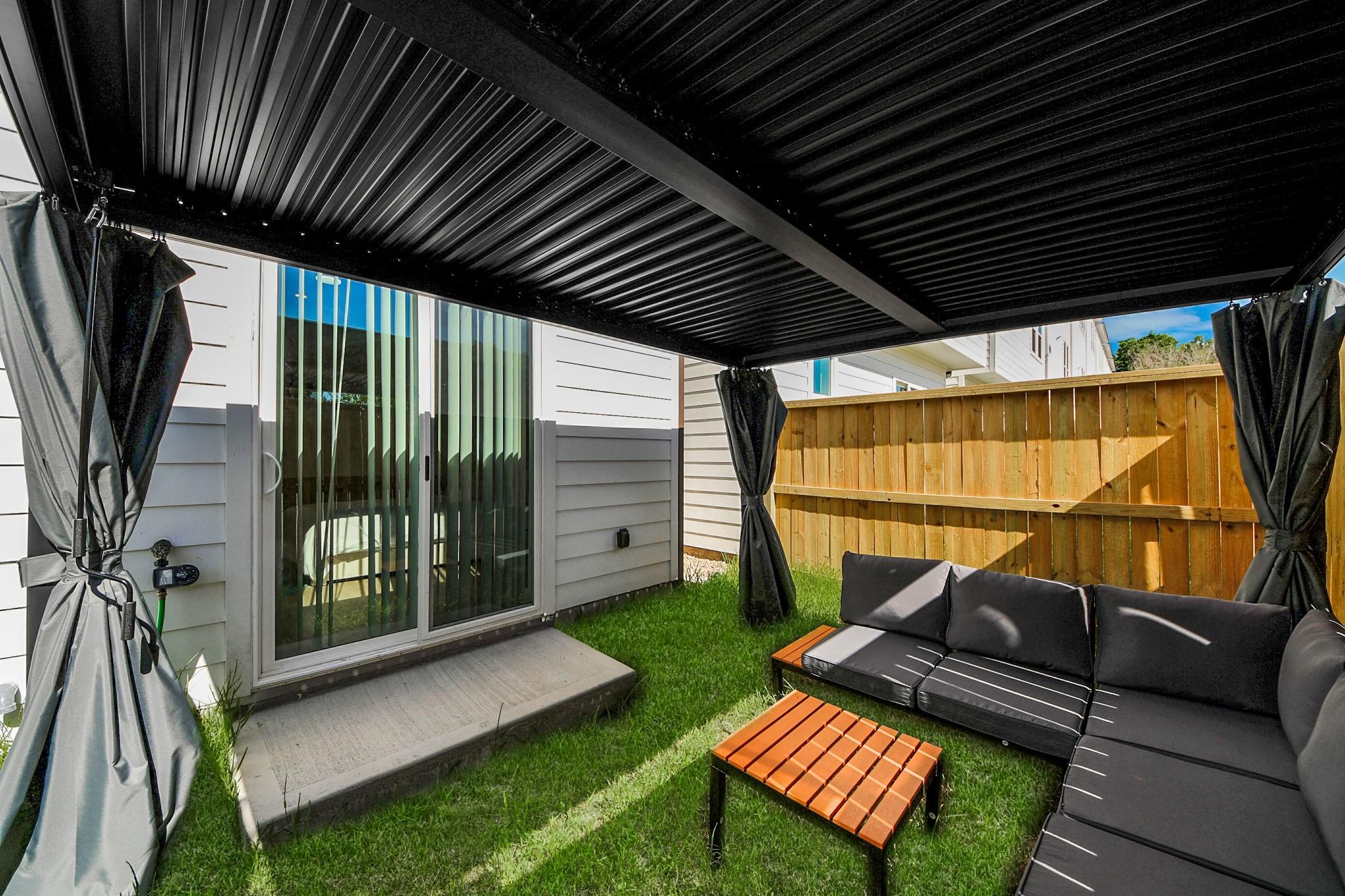 Patio with a black pergola, dark sectional sofa, wooden coffee table, and fenced grassy yard.