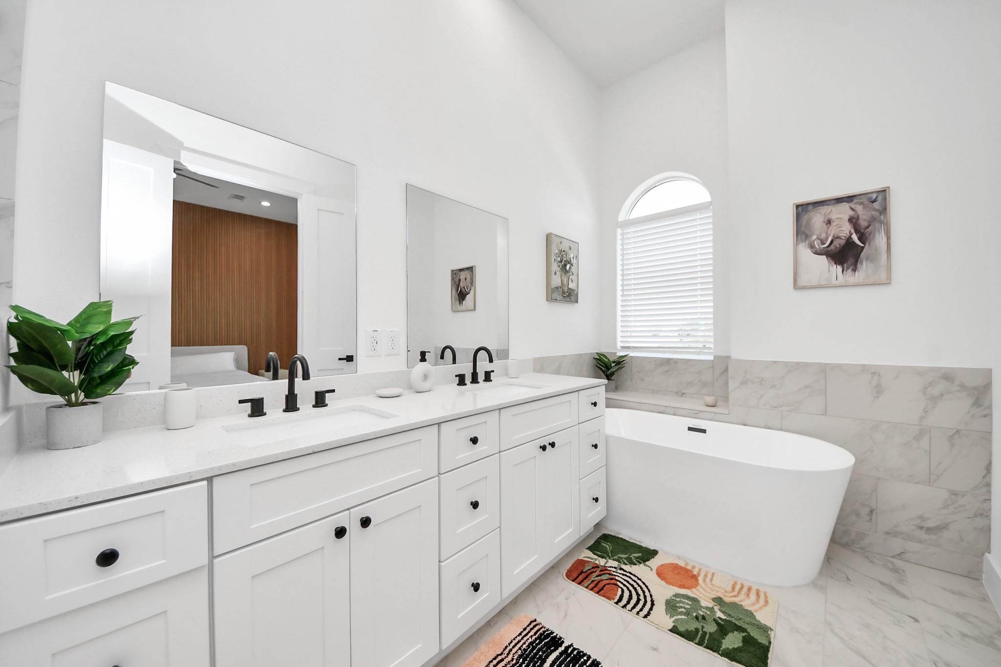 Bright bathroom with a double vanity and freestanding soaking tub beneath an arched window.