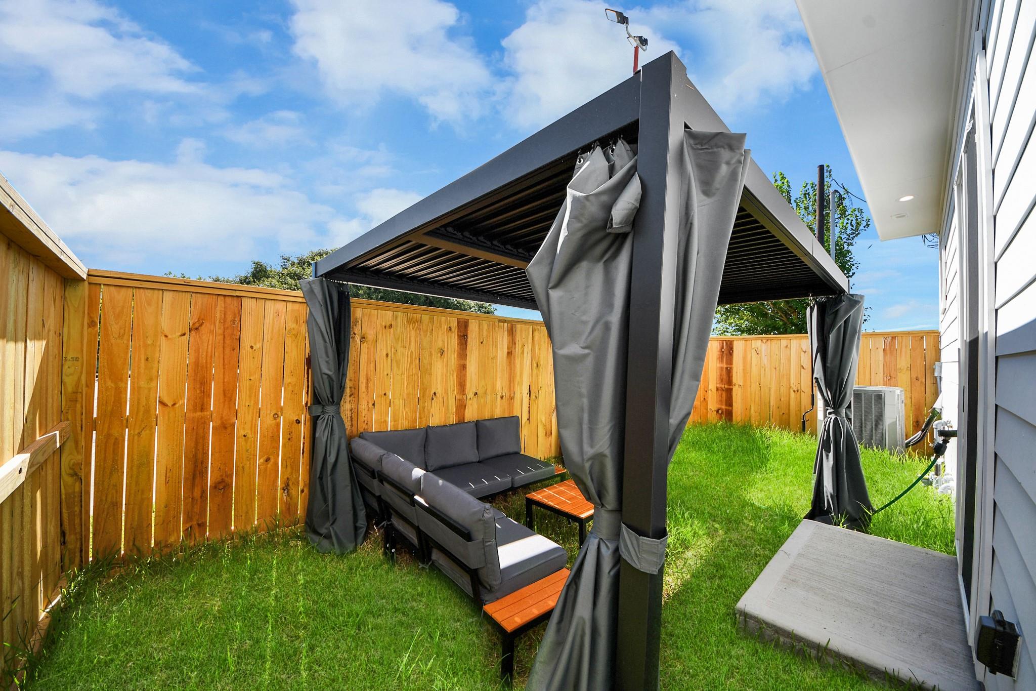 Fenced backyard with a curtained pergola, sectional seating, and green lawn.