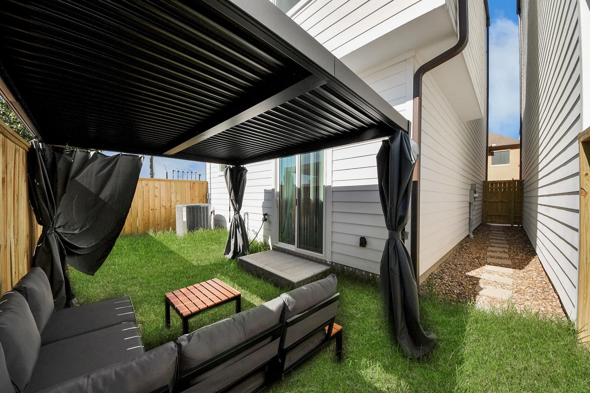 Fenced backyard patio with a black pergola, dark curtains, outdoor seating, and a gravel side path.