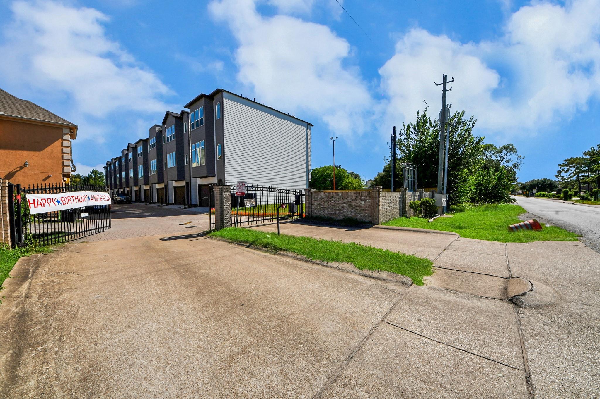 Gated row of modern townhomes with a paved driveway and a birthday banner on the entrance gate.
