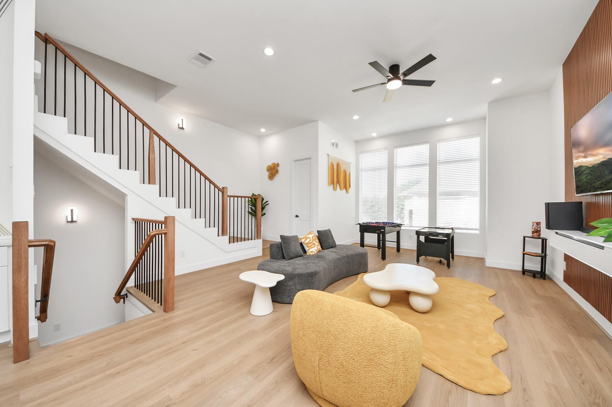 Staged living room with a modern wood-and-black staircase, gray sofa, and sculptural tables on a mustard rug.