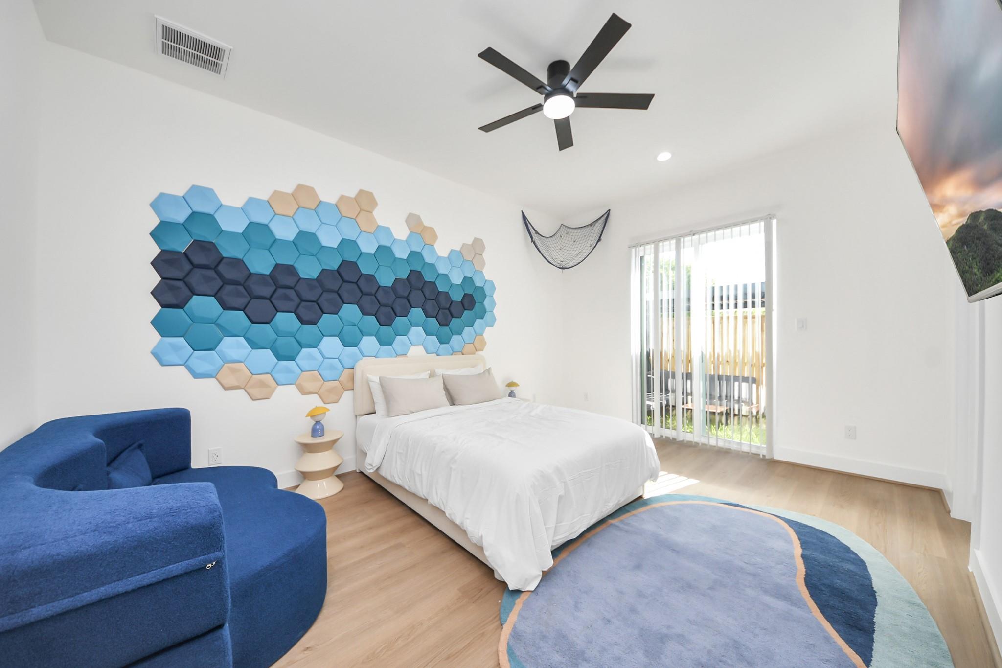 Staged bedroom with a white bed, blue sofa, and colorful hexagon wall art.