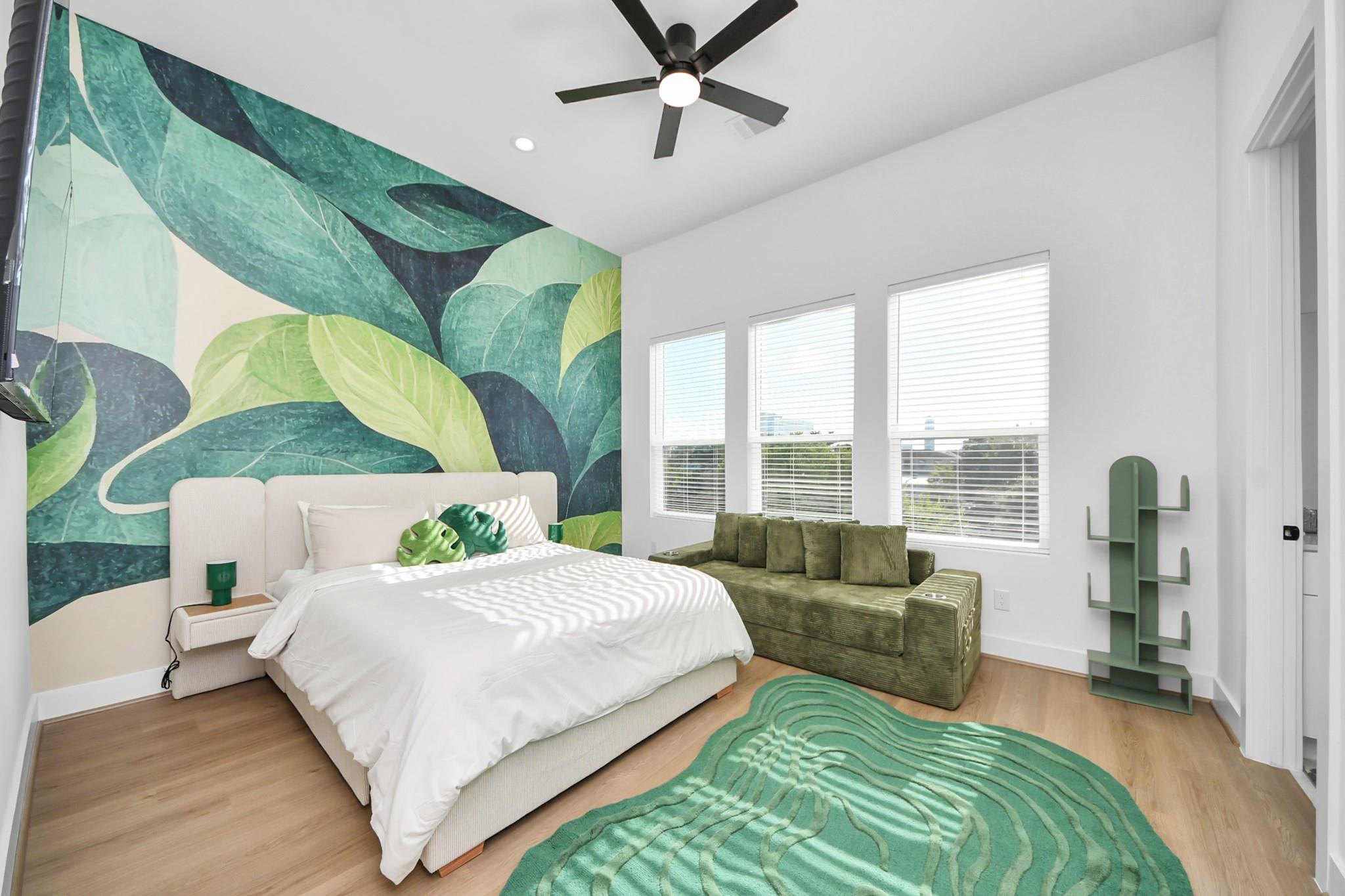Bedroom with a bold leaf mural, white bed, green sofa, and three bright windows.