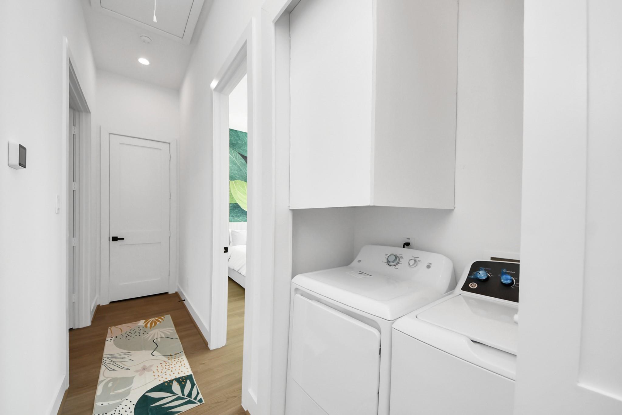 Laundry area with washer and dryer beneath white cabinets beside a hallway with a patterned runner.