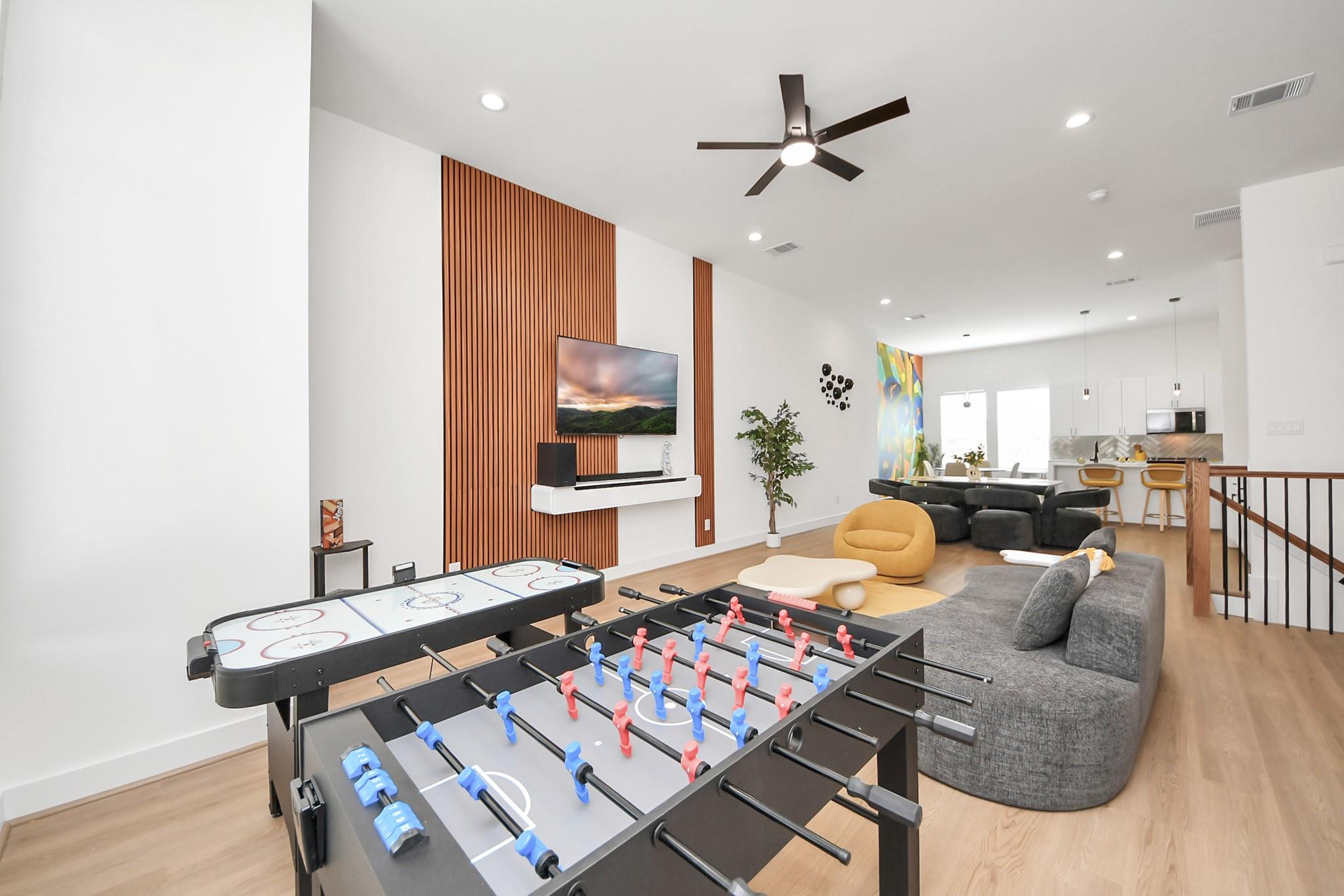Open-plan game room with foosball and air hockey tables, a gray sectional, and a wood-slat TV wall.