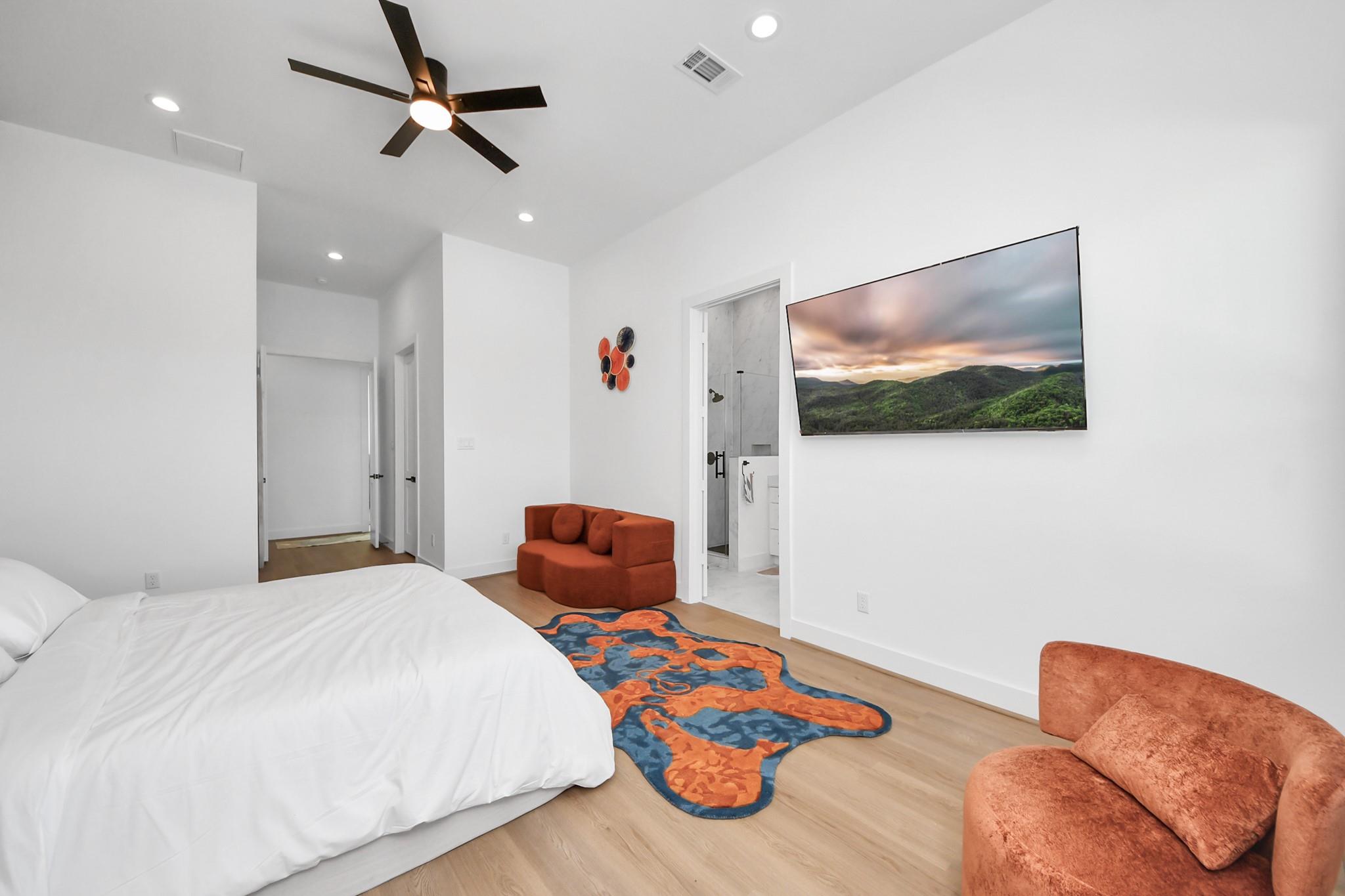 Bedroom with a white bed, colorful rug, rust-colored seating, and an open ensuite bathroom doorway.