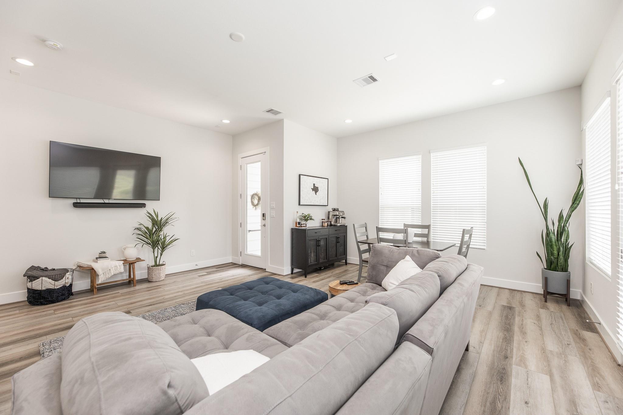 Furnished living room with a gray sectional, blue ottoman, and dining area by bright windows.