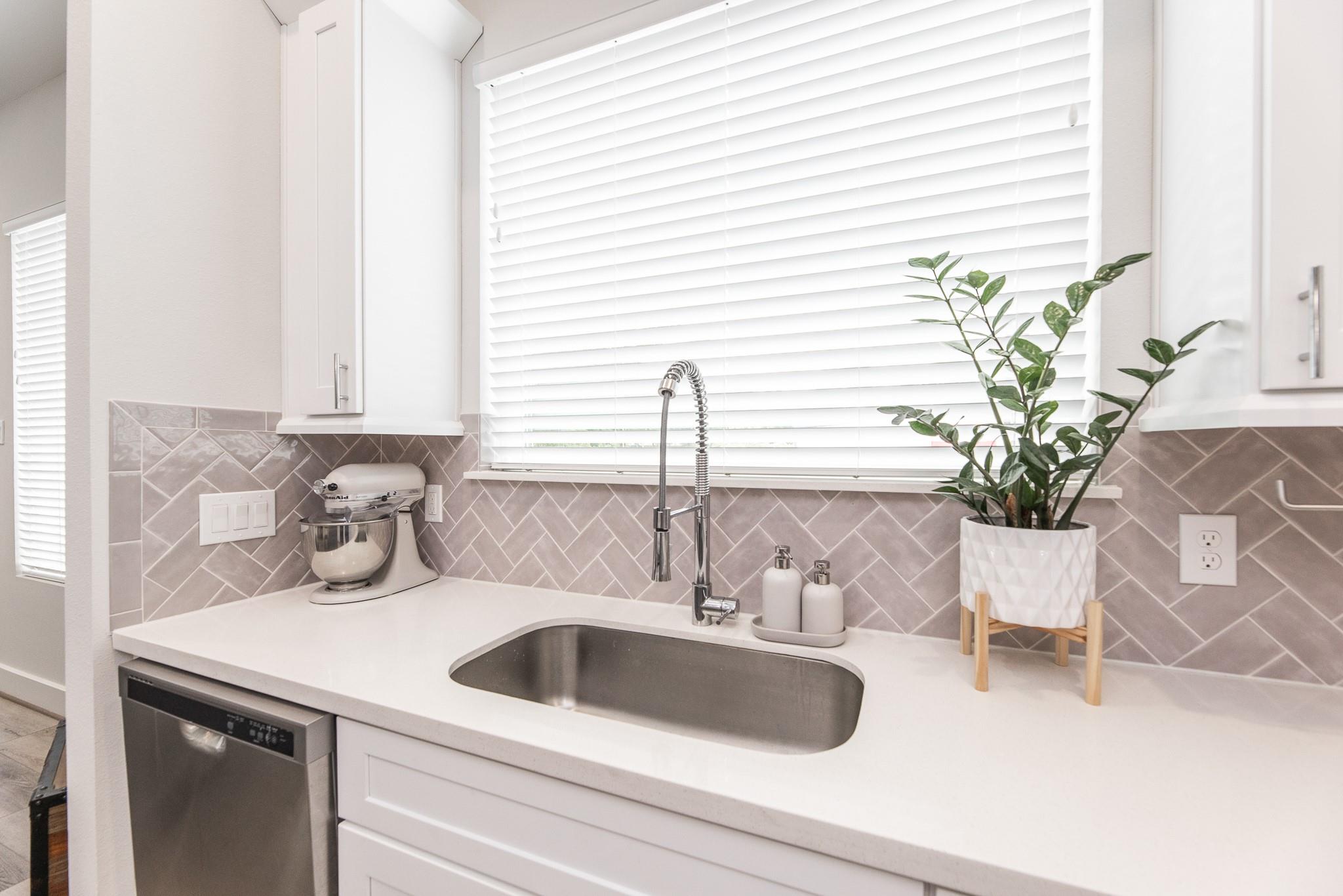 Kitchen sink beneath a wide window, with taupe geometric tile backsplash and white cabinets.