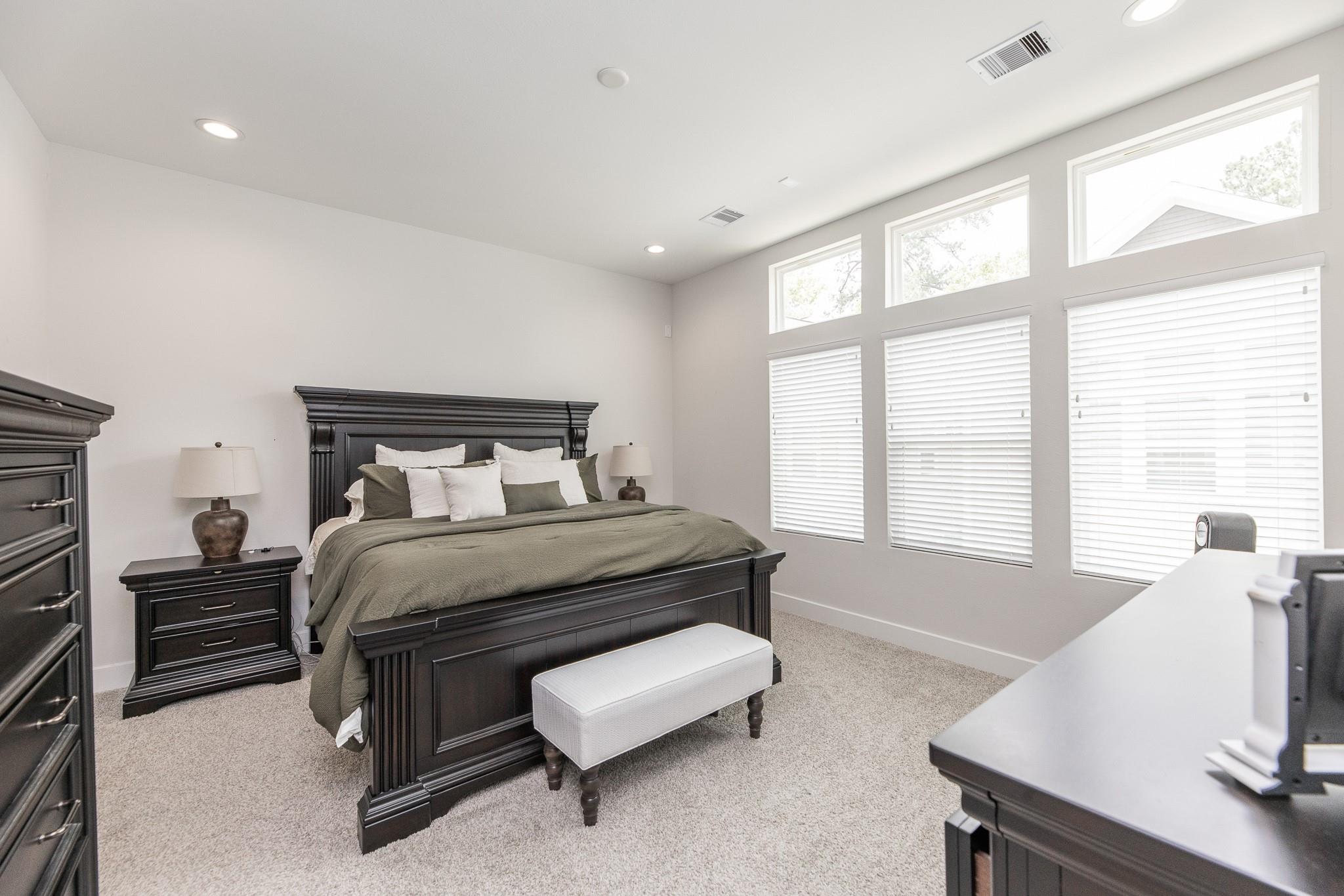 Furnished bedroom with a dark wood bed, gray bedding, and tall windows with blinds.